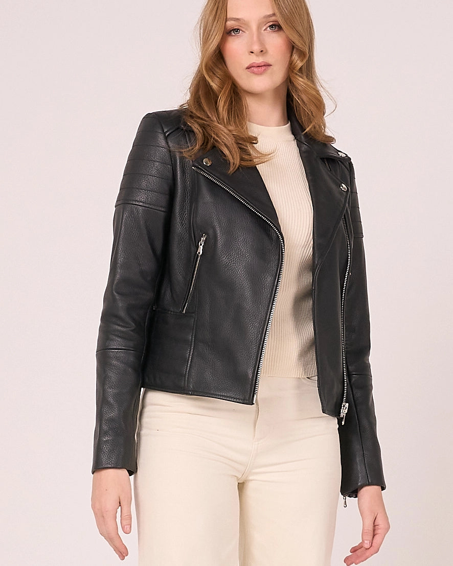 A woman wearing a black real leather moto jacket over a cream ribbed top and cream pants, standing against a solid light beige background.