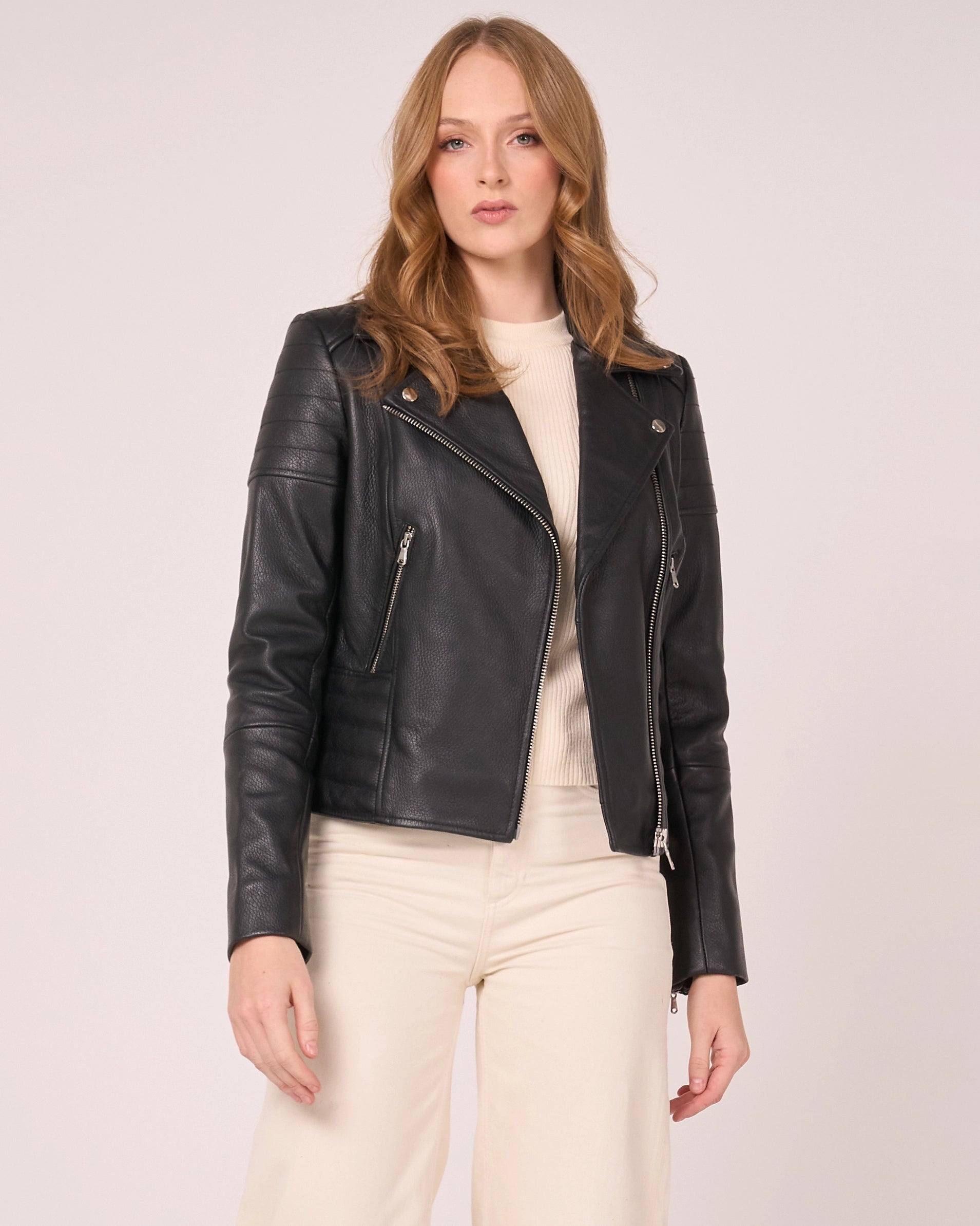 A woman models a black leather moto jacket with silver zippers and snap details, worn over a cream top and cream pants, against a light grey studio background.