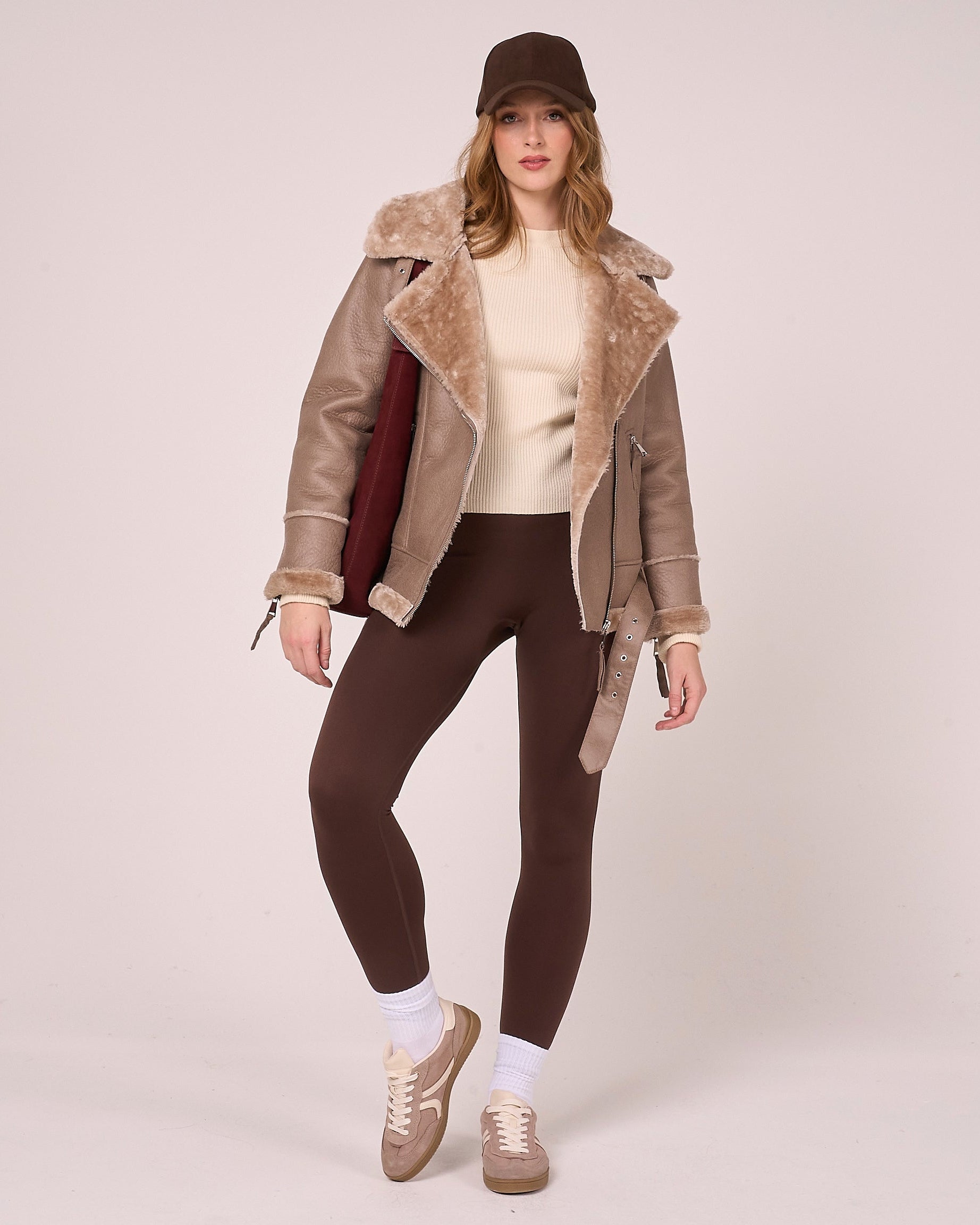 Woman styles faux shearling aviator coat with cream sweater, brown leggings, socks and trainers. The look is finished with a brown cap and burgundy suede bag.