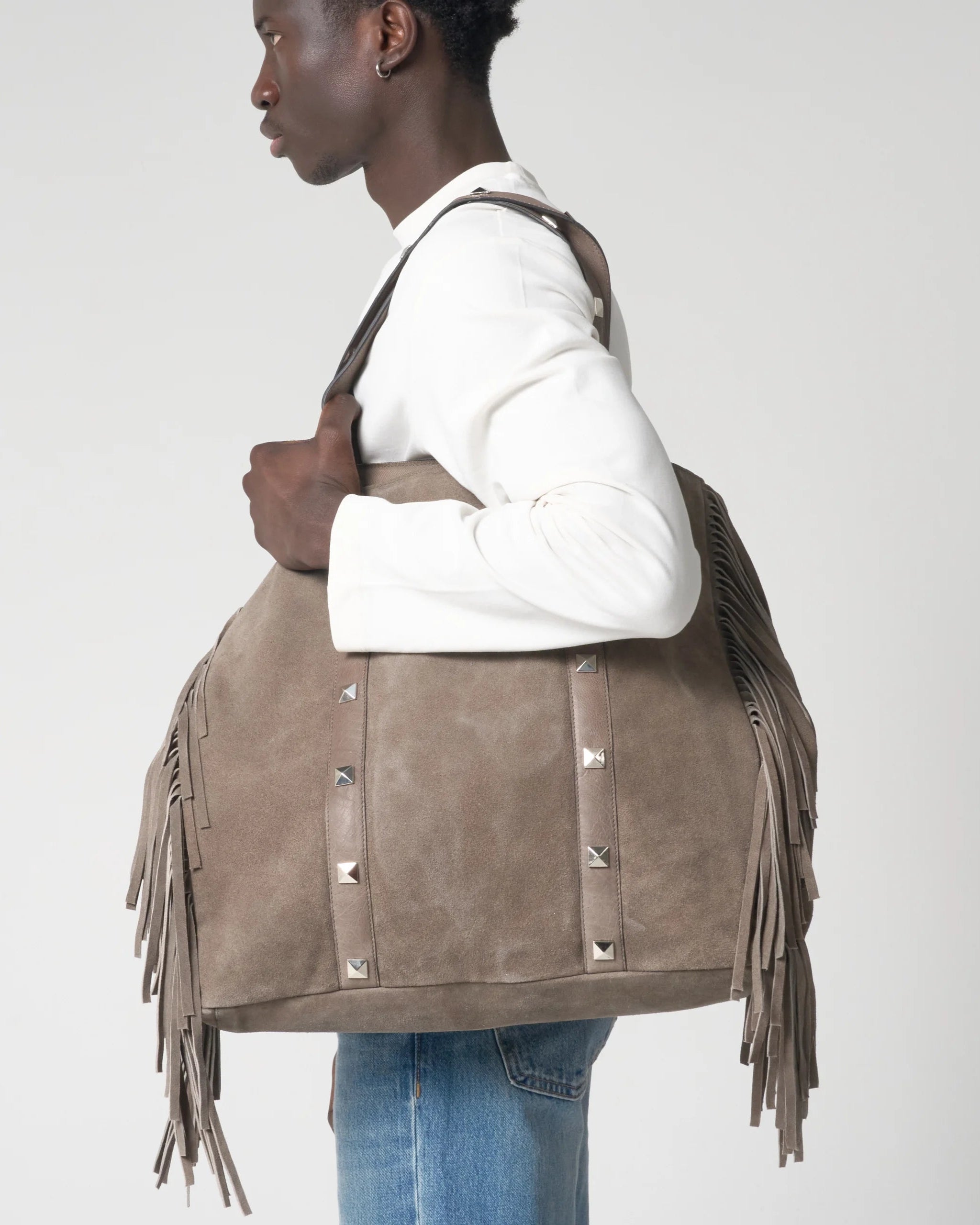 Person holding a brown suede handbag with fringe and studs on a white background