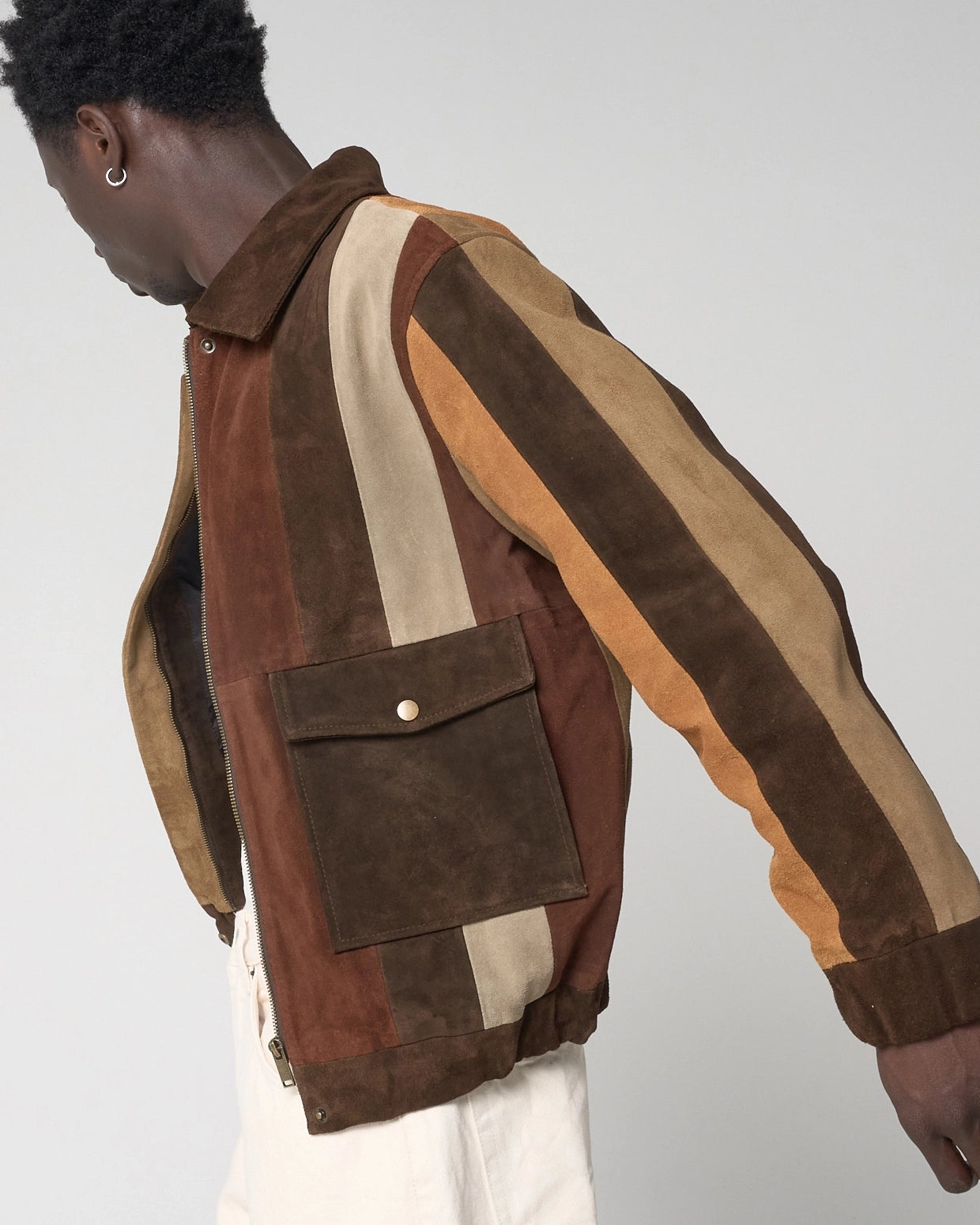 A man wearing a brown suede patchwork jacket with vertical stripes in dark brown, tan, and beige. The man stretched out his arm to display the stripes in full. The front of the jacket also has dark brown patch pockets.