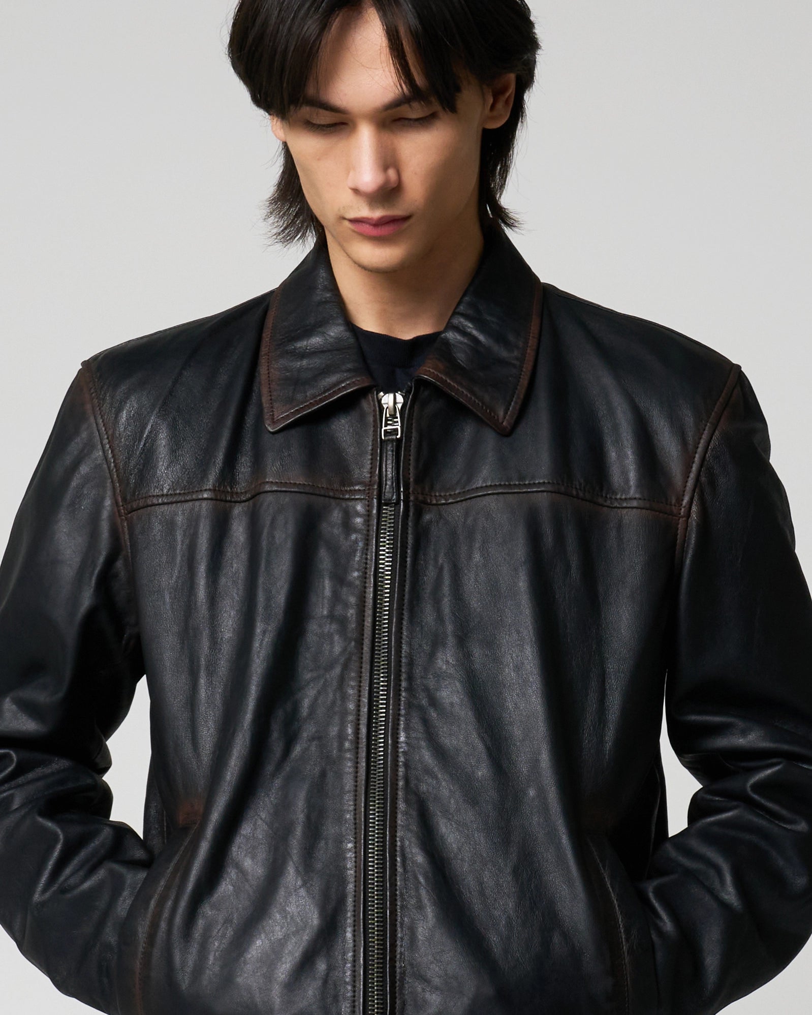 A man wearing a black leather jacket with brown contrast washed detail, creating a 'rust effect'. The jacket is fully zipped and the man has his hands in his pockets.