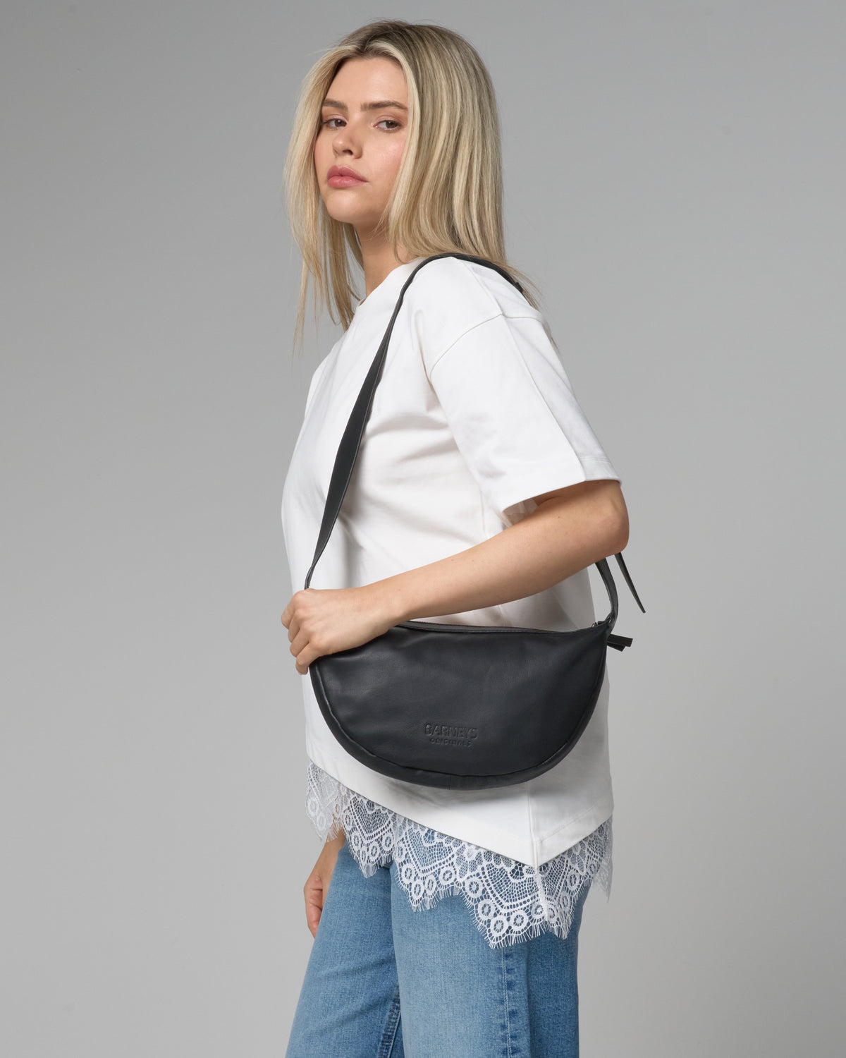 A blonde woman wearing a white t-shirt with lace hem and blue jeans, carrying a black crescent-shaped shoulder bag with a black strap against a gray studio background.