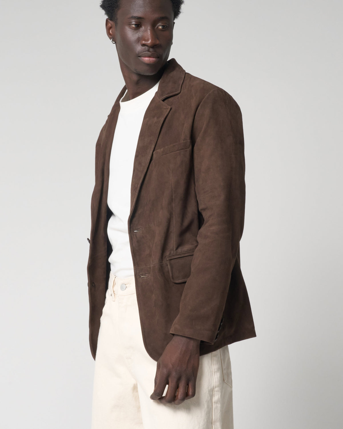 A man wearing a brown suede blazer over a white t-shirt and cream-colored trousers, standing against a light grey studio background.
