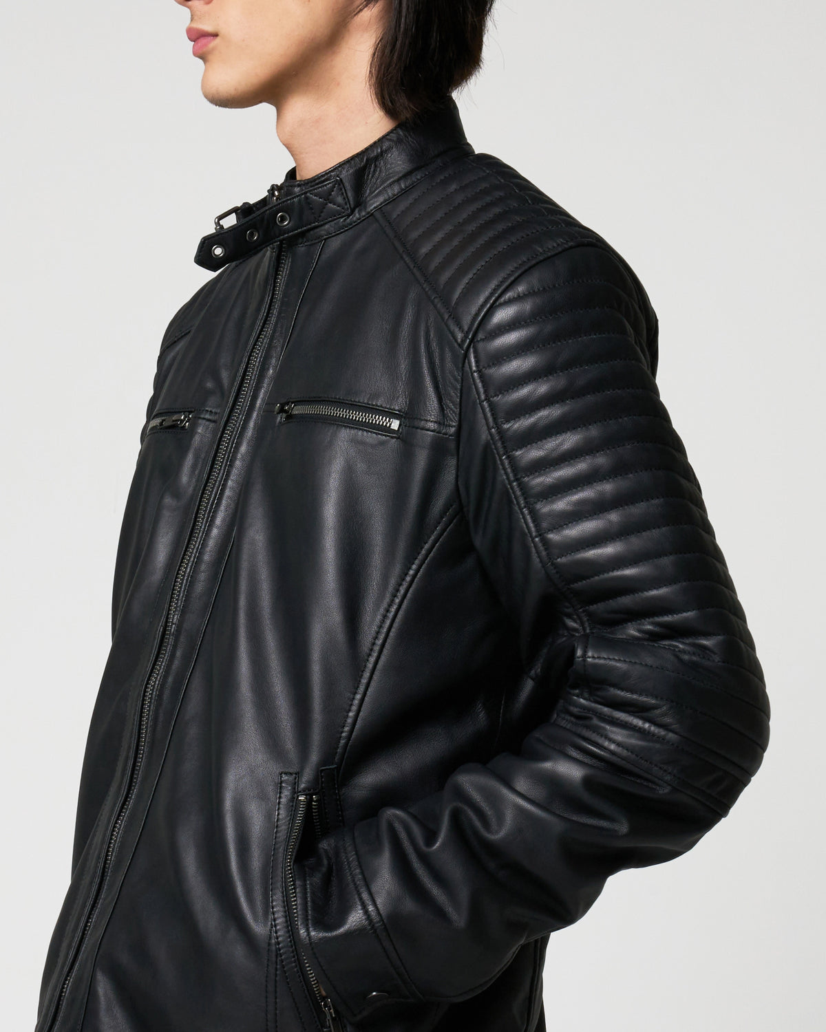 A close-up side view of a person wearing a black leather jacket with quilted shoulder panels and zippered chest pockets, photographed against a light grey studio background. The neckline has a buckle on the neckline.