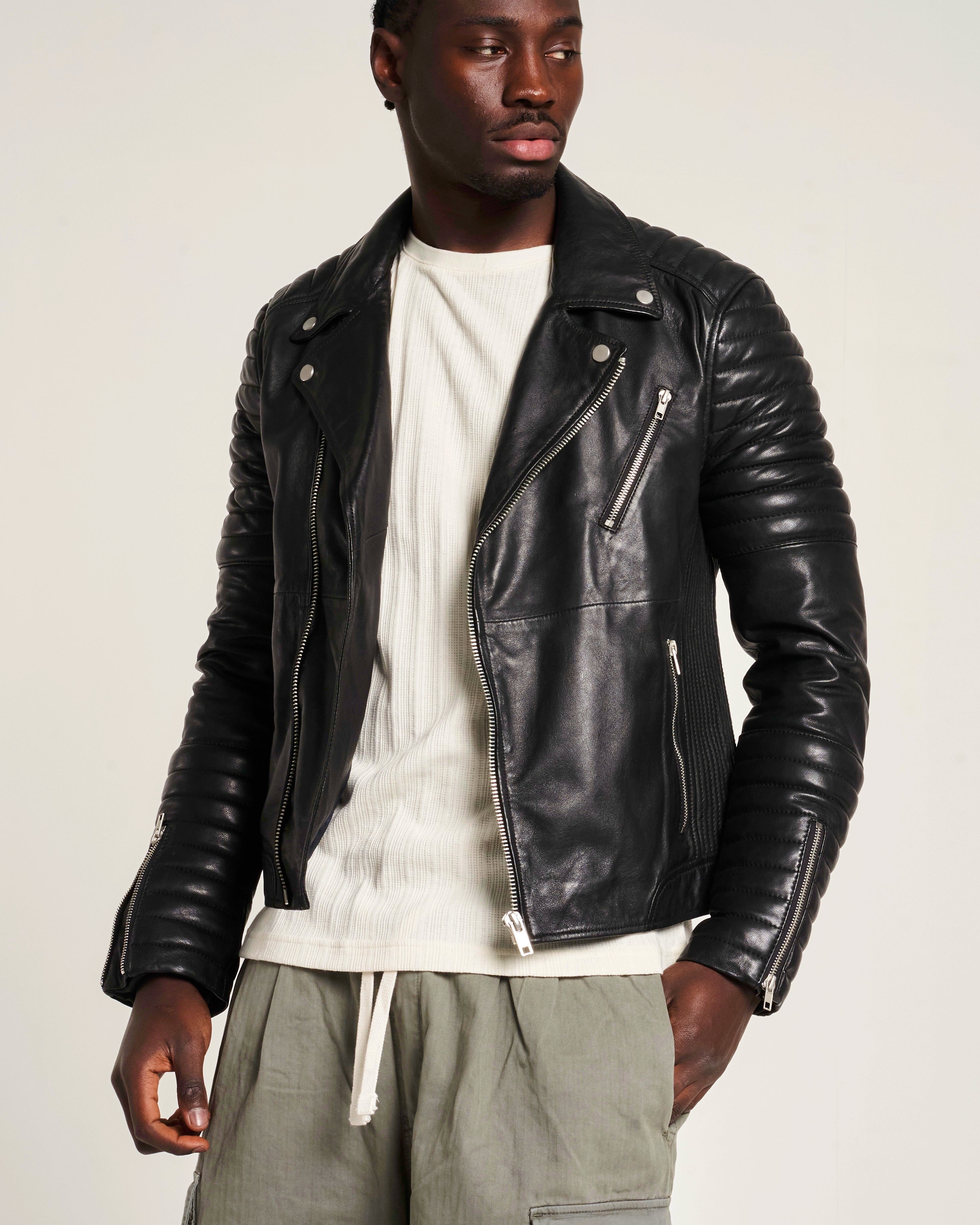 A man wearing a black leather biker jacket over a cream ribbed knit sweater and olive green drawstring pants. The sleeves on the jacket have padded ribbed detail.