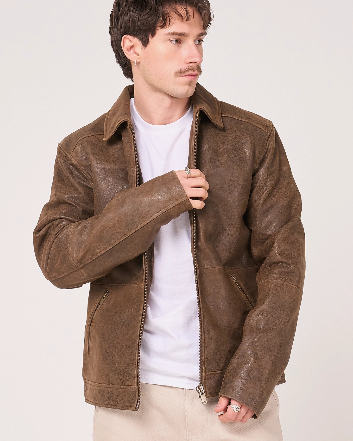 A man wearing a brown leather jacket over a white t-shirt and beige pants, standing against a light grey studio background.