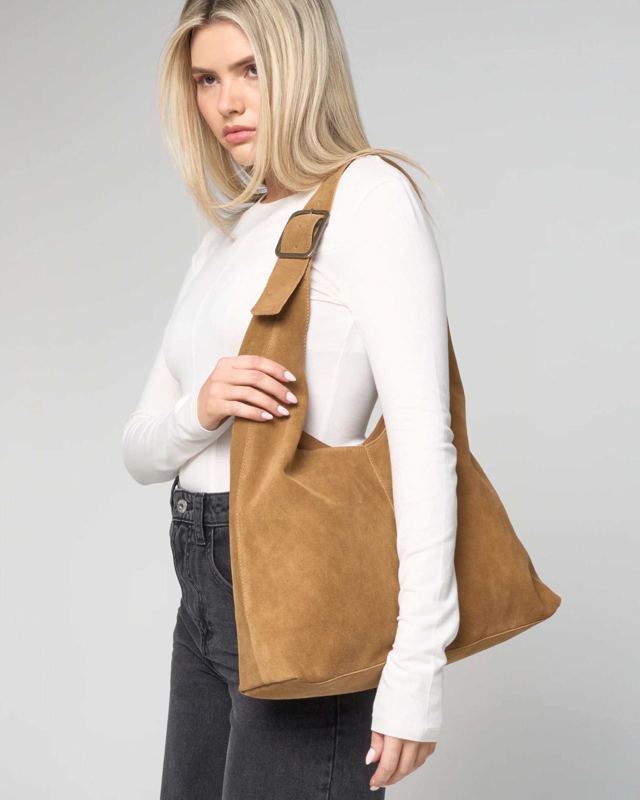 Image shows a model wearing the tan suede slouch bag from Barneys Originals, with a white long sleeve tshirt and grey jeans.