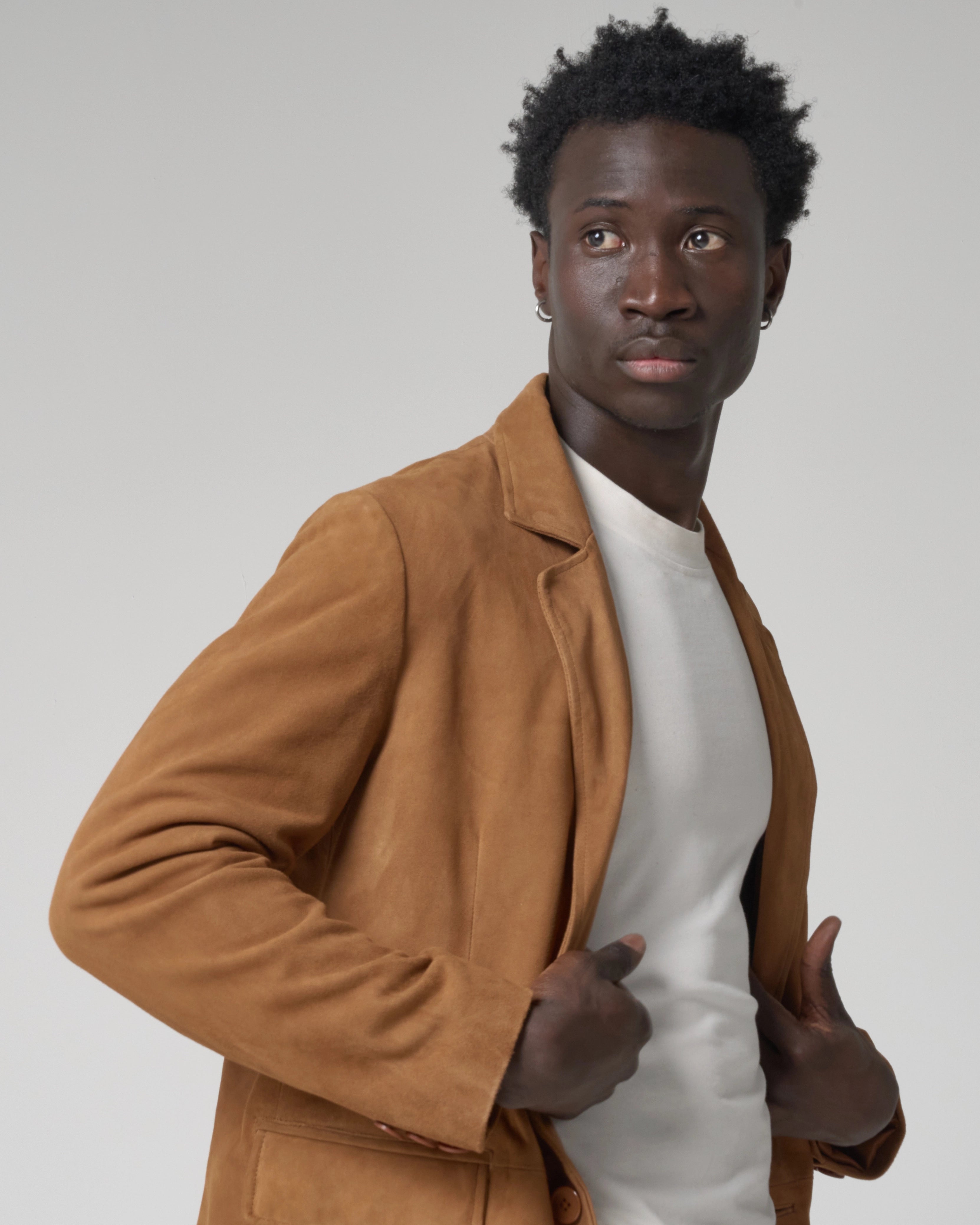 A man wearing a tan brown suede blazer over a white crewneck t-shirt. The man holds the lapels of the blazer jacket open.