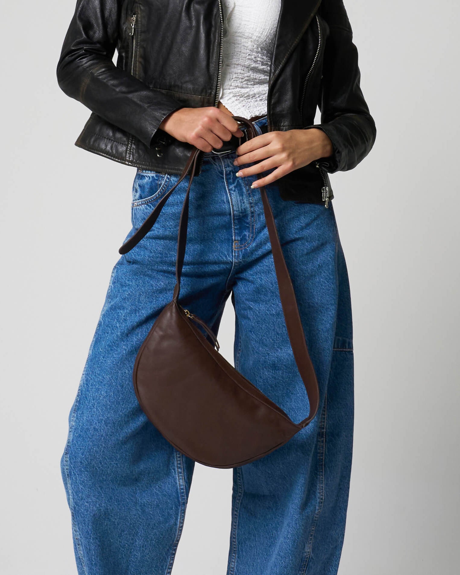 Image shows a brown cross body bag made from leather, held in the hands of a woman wearing a leather jacket, white tee and jeans.