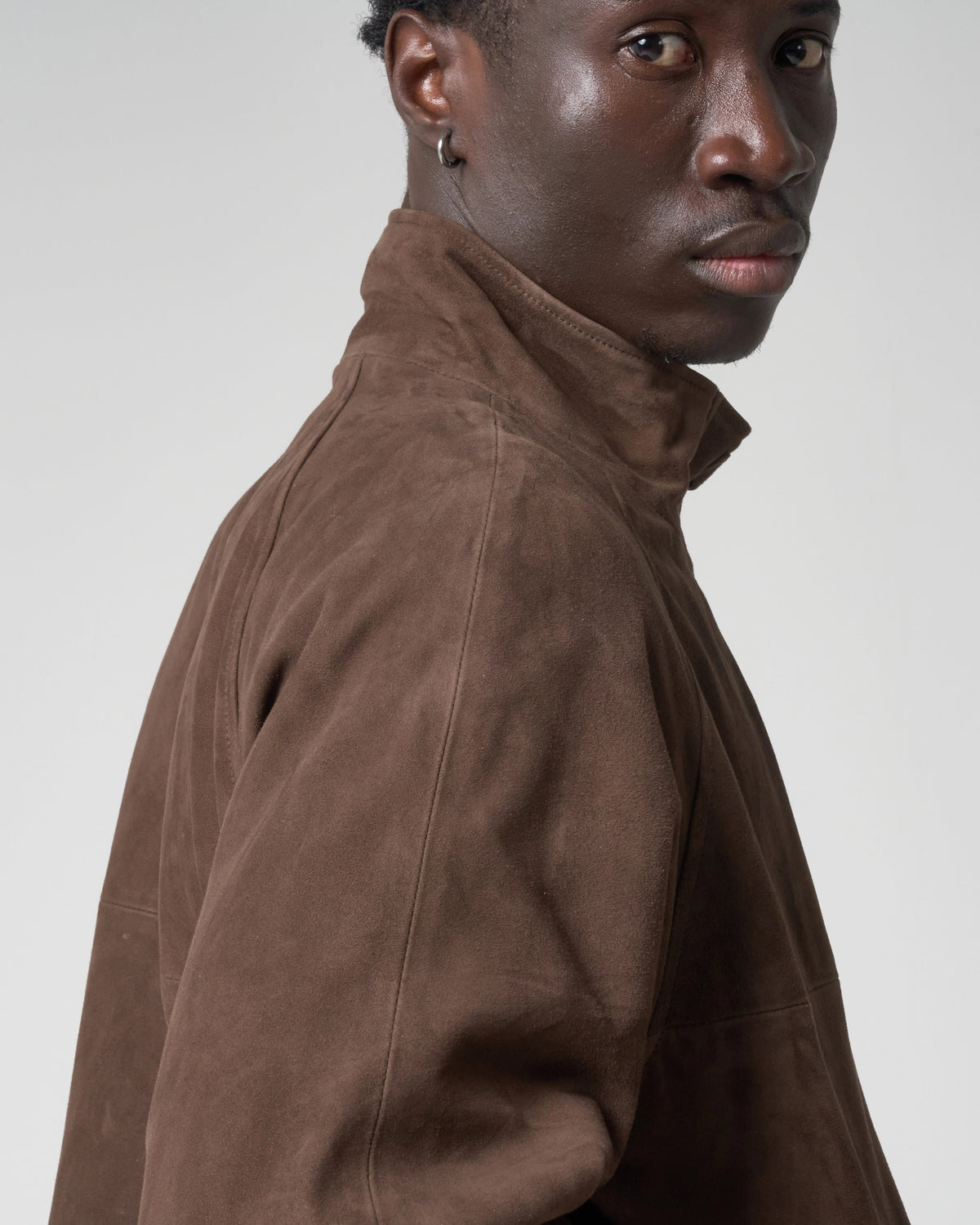 A close-up side profile of a model wearing a brown suede jacket against a light gray studio background.