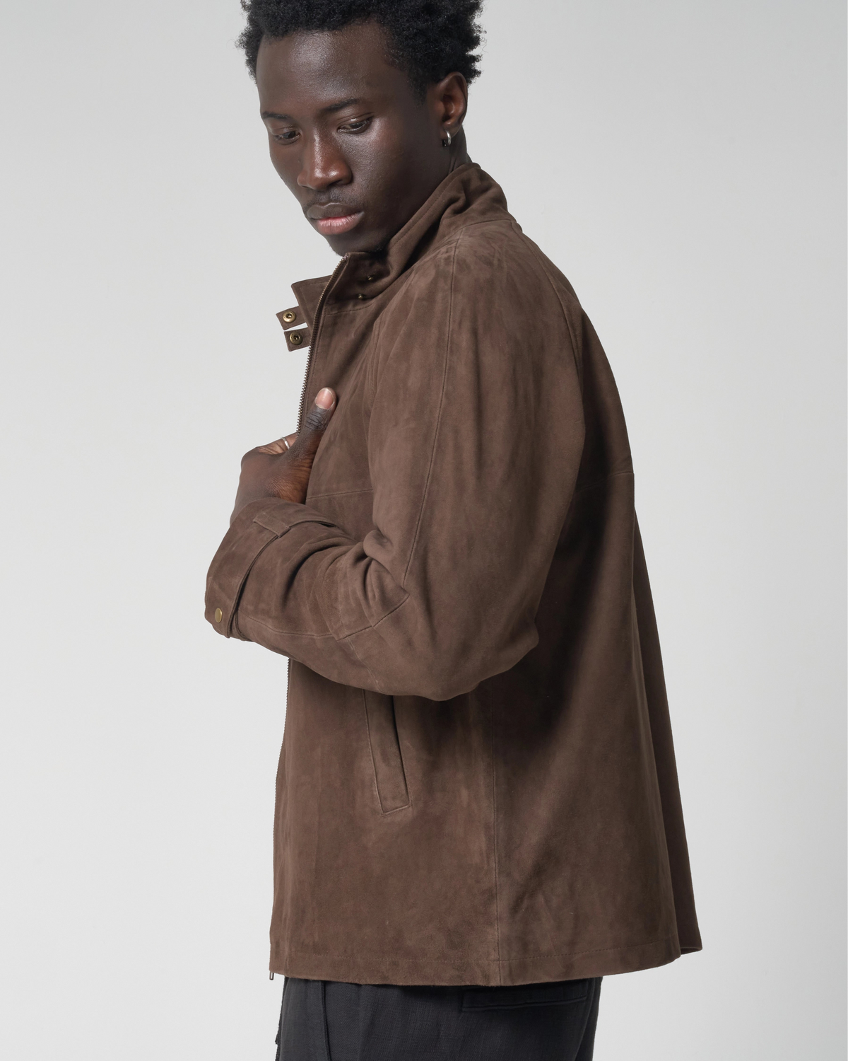 A man wearing a brown suede jacket, shown from a side profile against a light gray studio background.