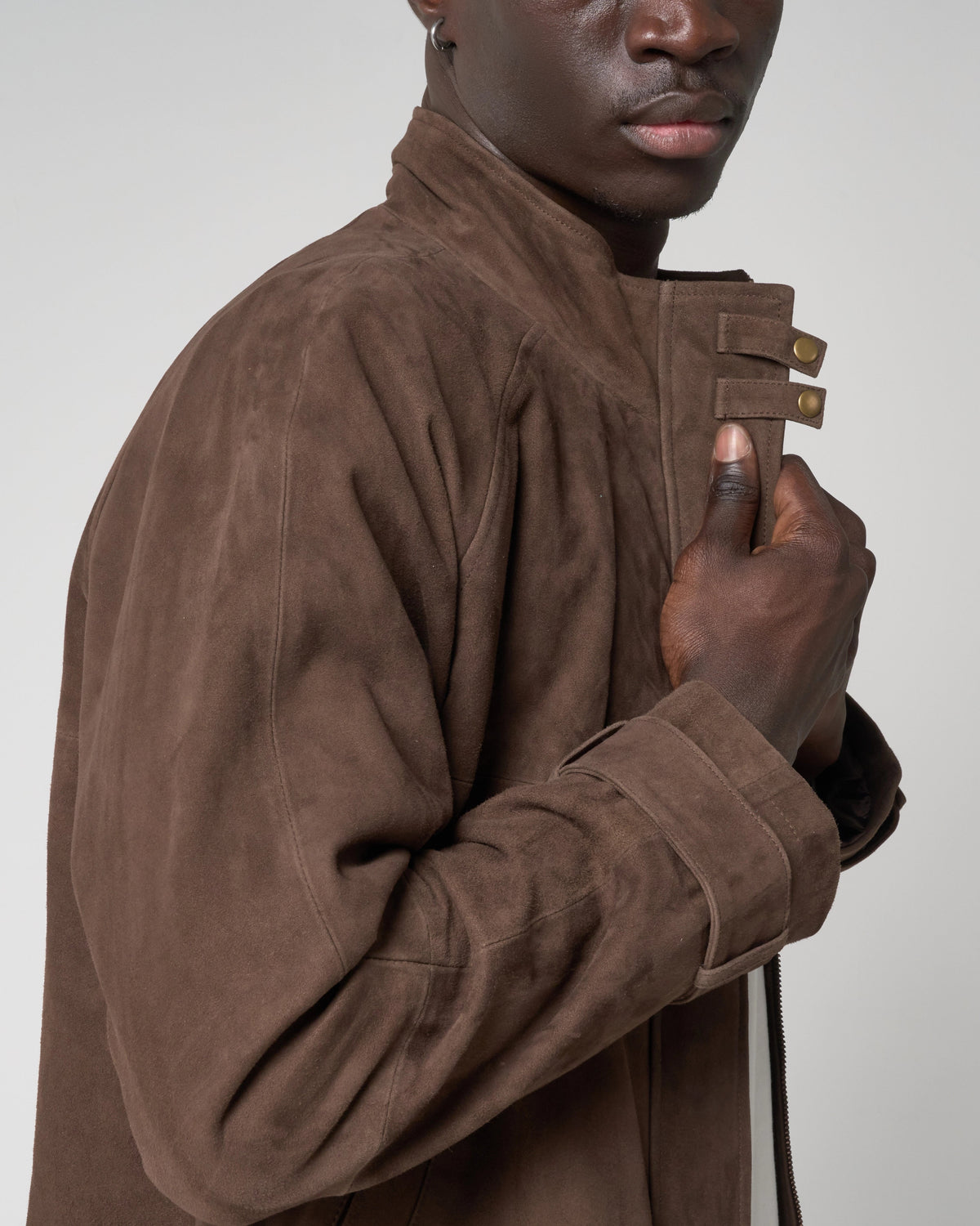 A close-up of a model wearing a brown suede jacket with a high collar and unique strap closures on the front, set against a plain light gray background.