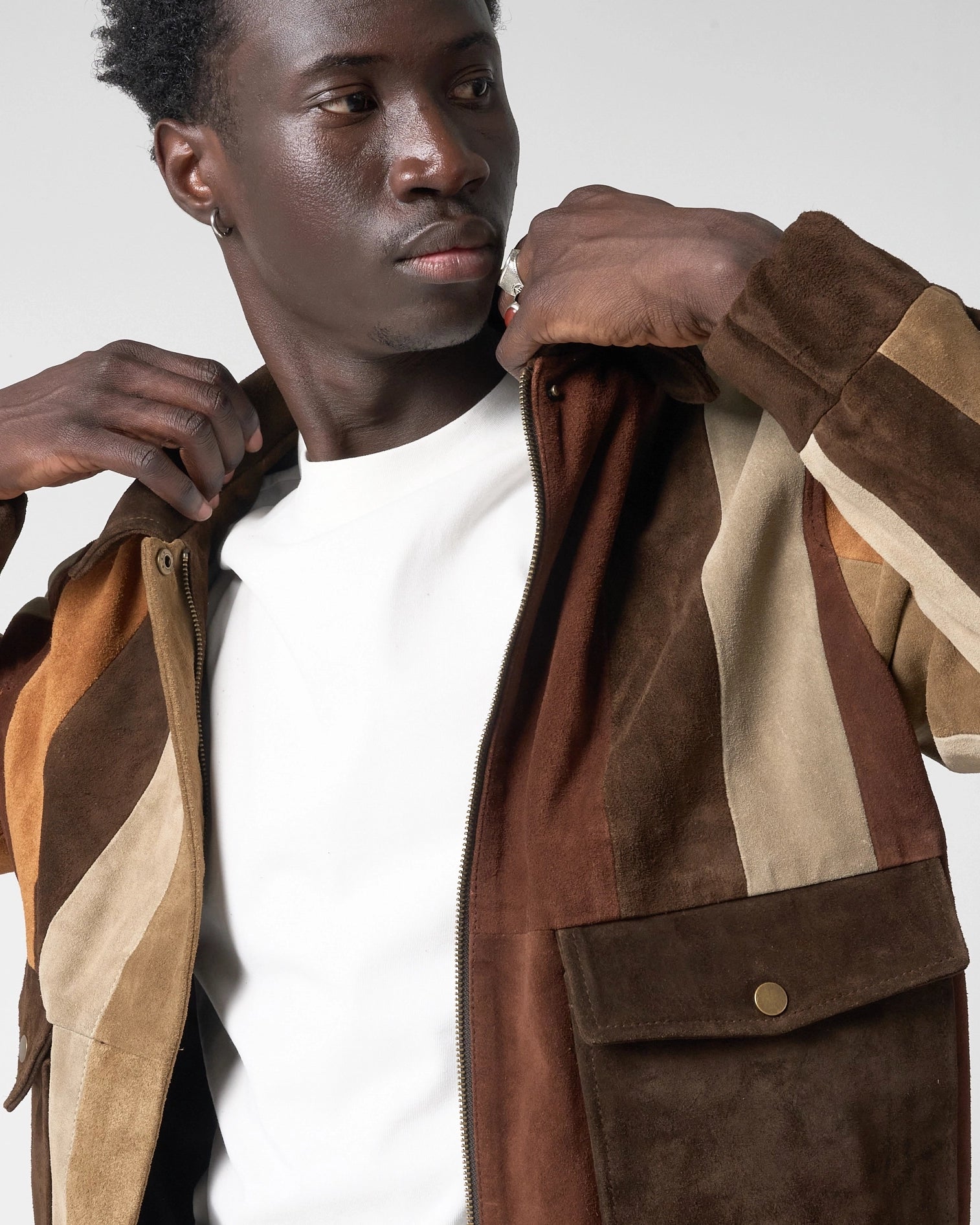 A close-up of a man wearing a brown buffalo suede zip-up bomber jacket with color-blocked panels in dark brown, tan, and beige over a white t-shirt.