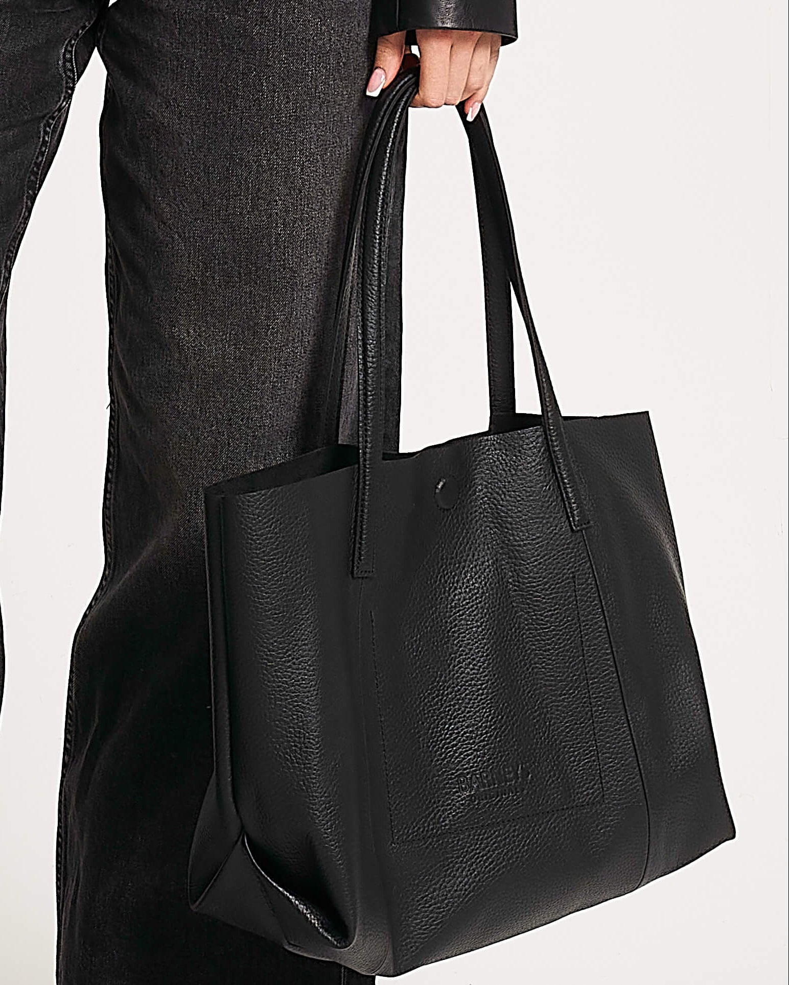 Person holding a black leather tote bag against a neutral background. The bag has two straps and is embossed with 'Barneys Originals'.