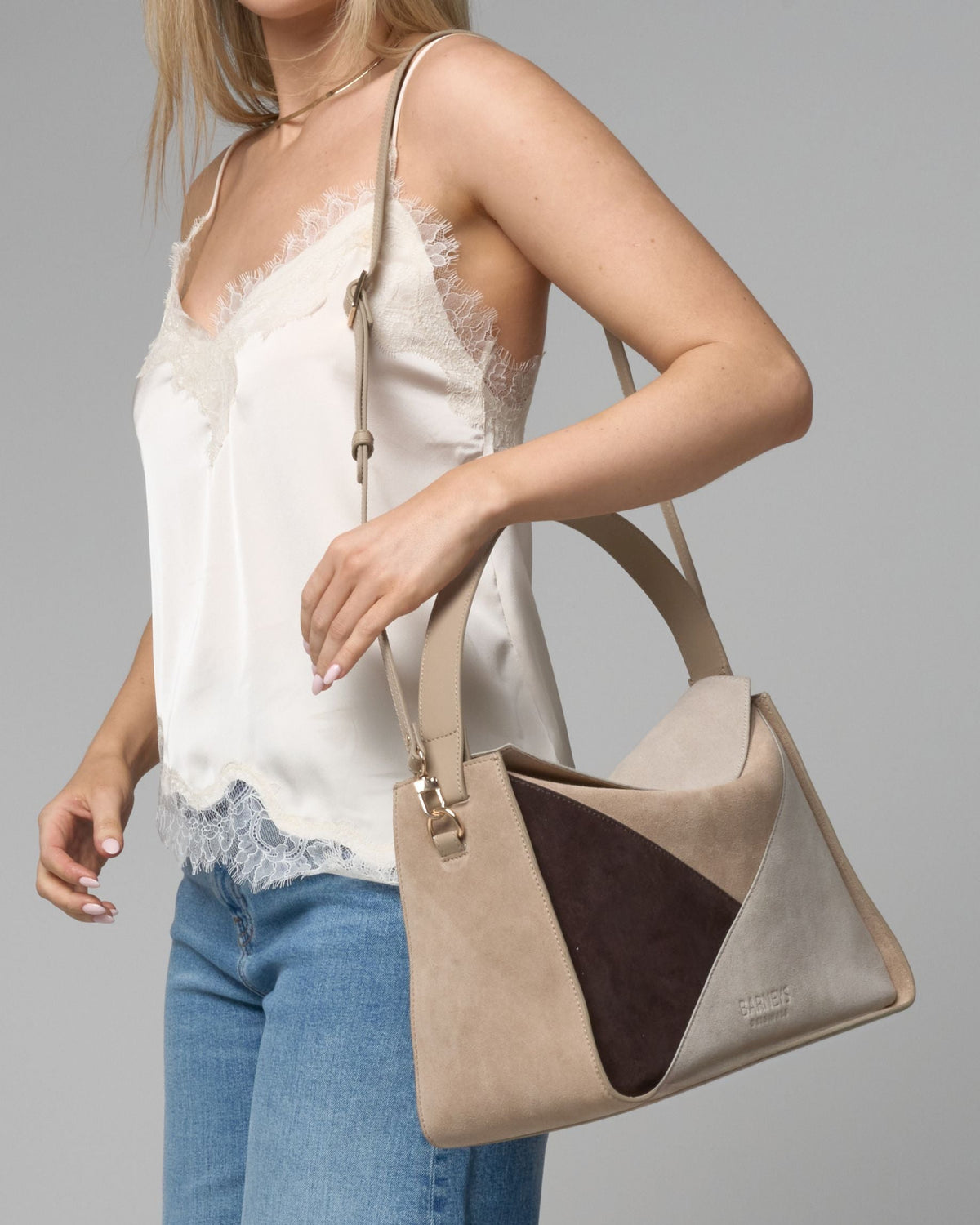 A woman wearing a white lace-trim camisole and blue jeans carries a structured beige handbag with a dark brown triangular accent panel and a light beige shoulder strap against a solid gray studio background.
