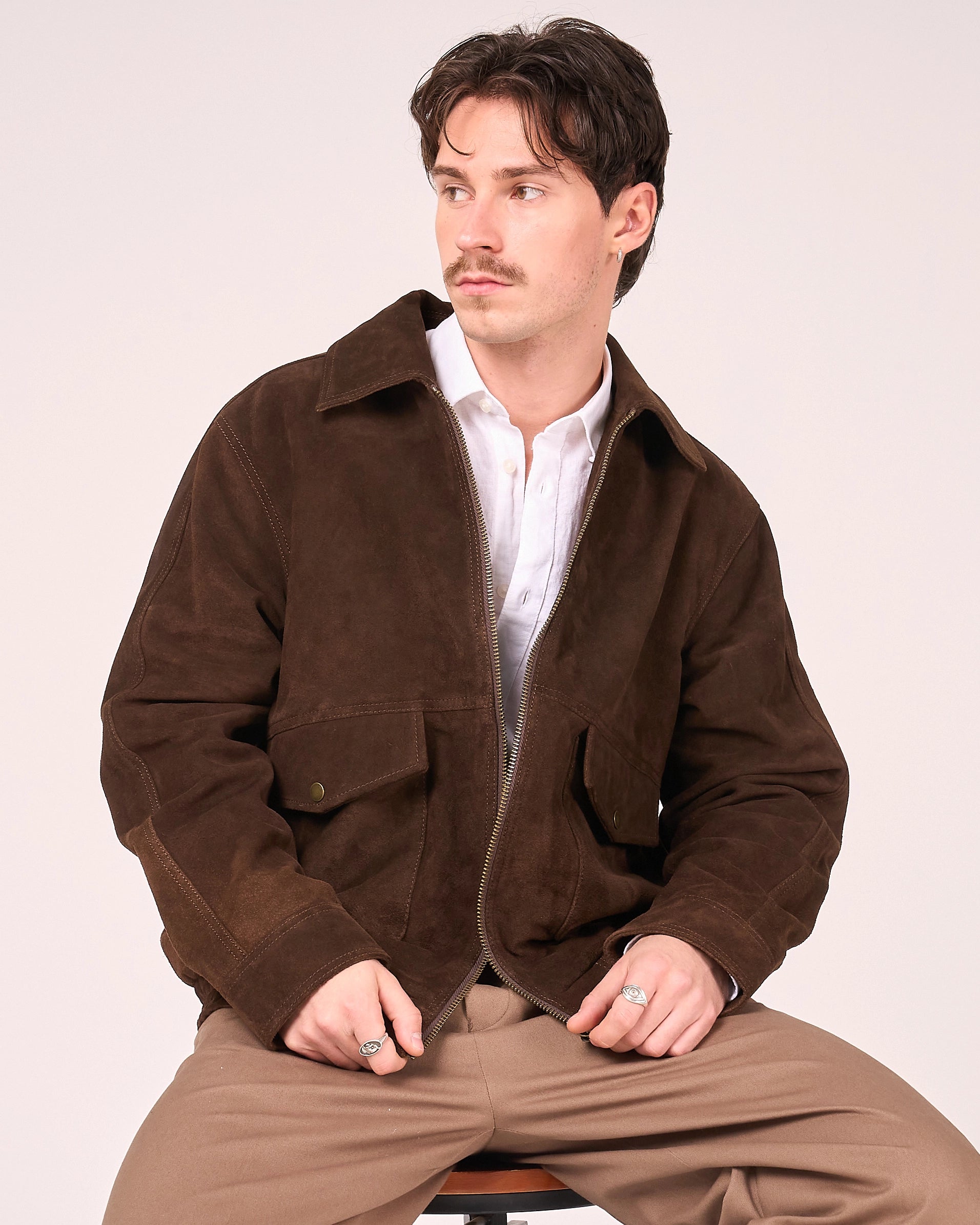 Man sitting in a photo studio whilst wearing the brown buffalo suede jacket by Barneys Originals. The jacket is unzipped and has two patch pockets on the lower waist.