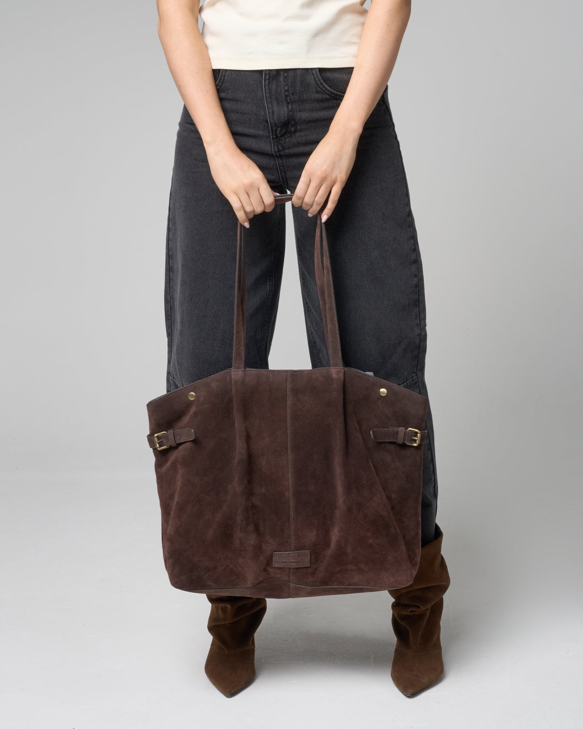 A person wearing dark jeans and brown boots holds a large dark brown suede tote bag with double shoulder straps and buckle details against a light gray studio background.