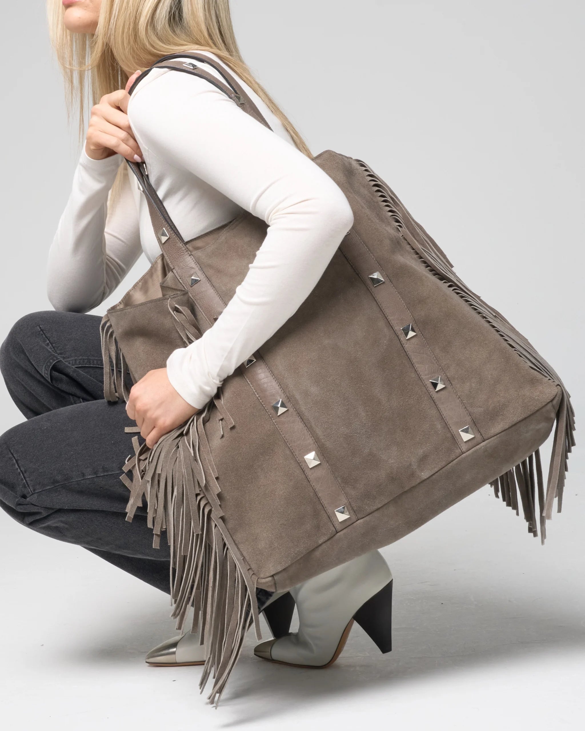 Woman holding a large grey suede tote bag with fringe details and studs.
