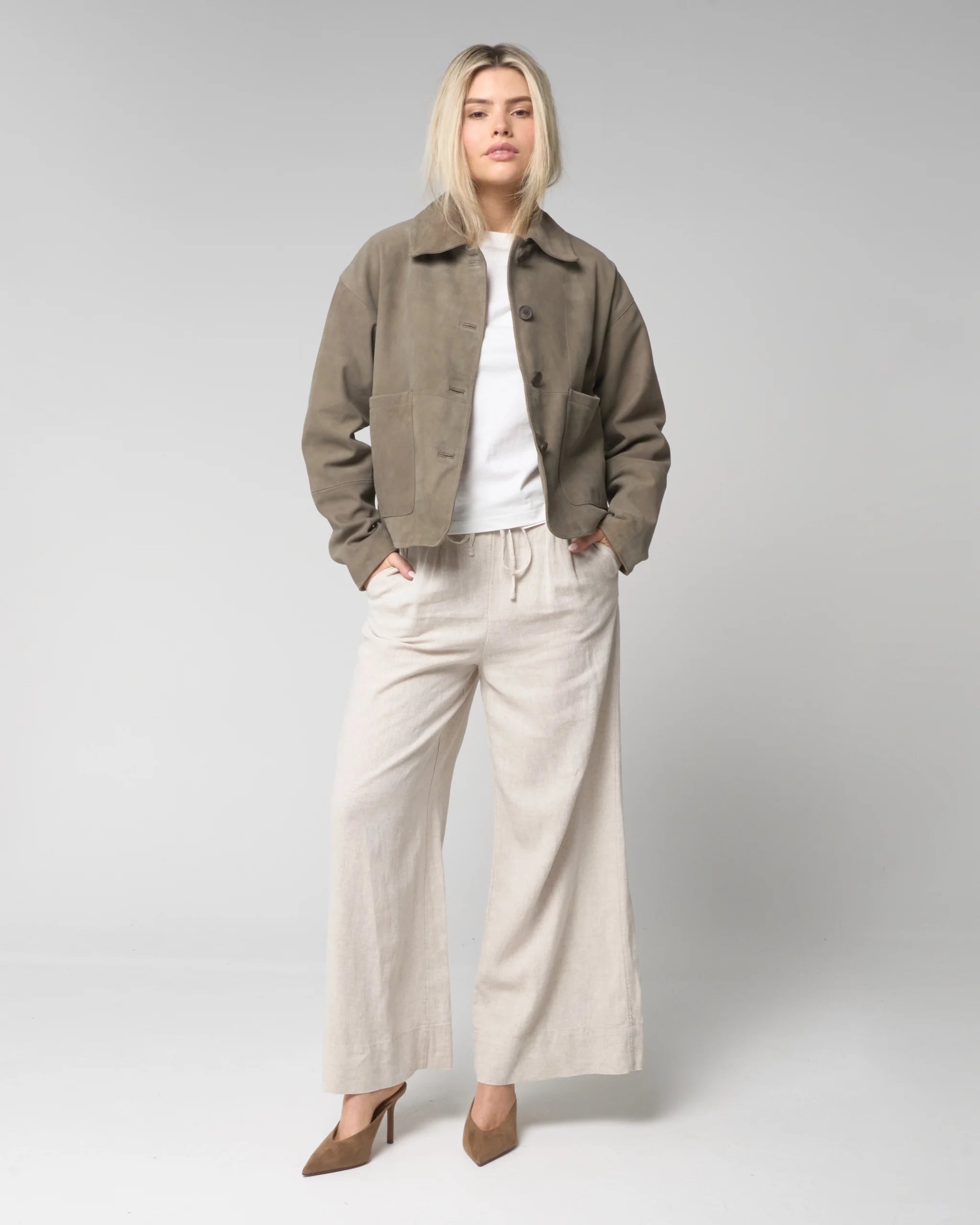 Full outfit image of woman wearing the sage suede jacket, unbuttoned and open to expose the outfit below. The sage suede jacket is cropped at the waist and the woman wears the item with linen trousers and heels.