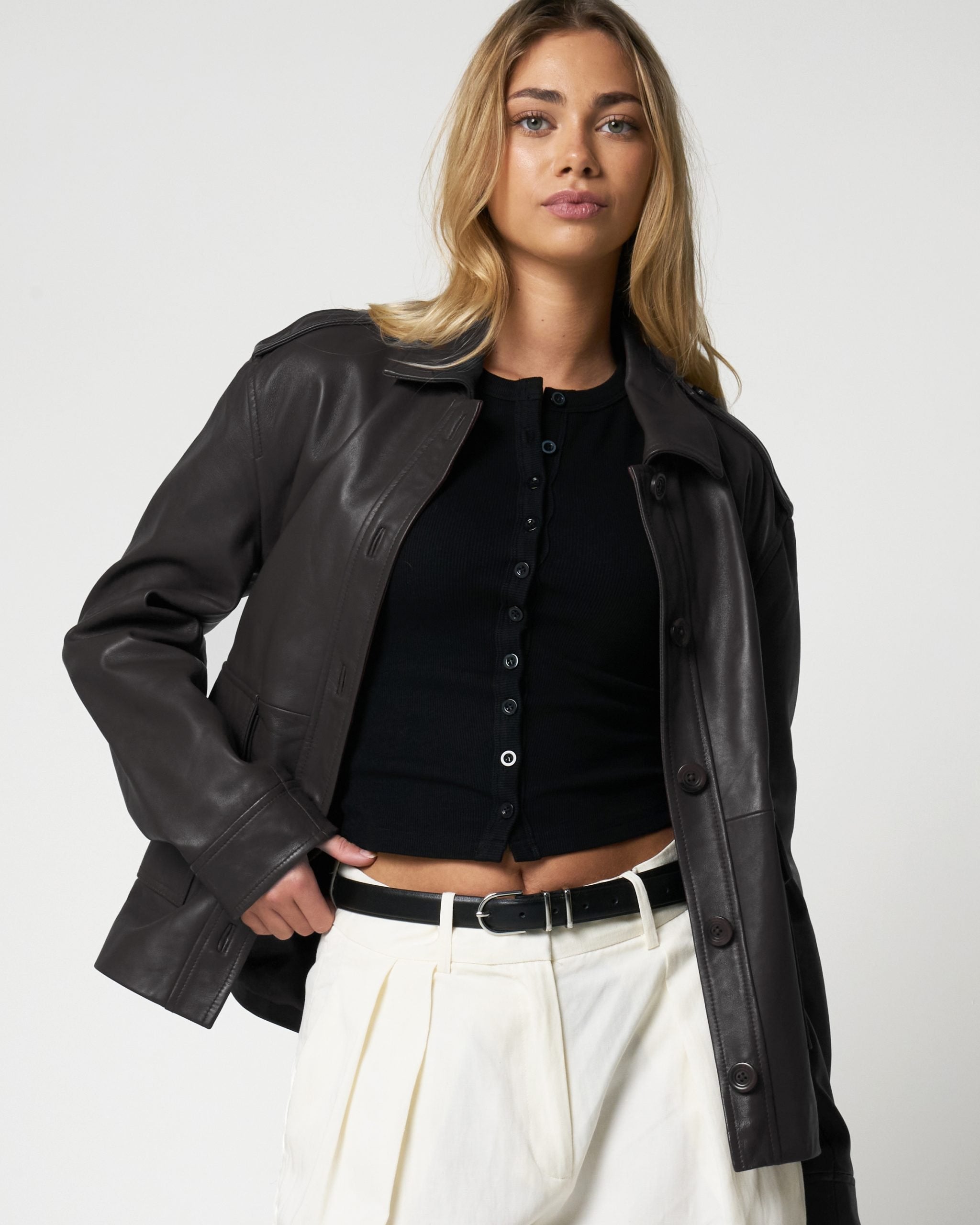 Woman wearing a black leather jacket over a black shirt and white pants on a light gray background.