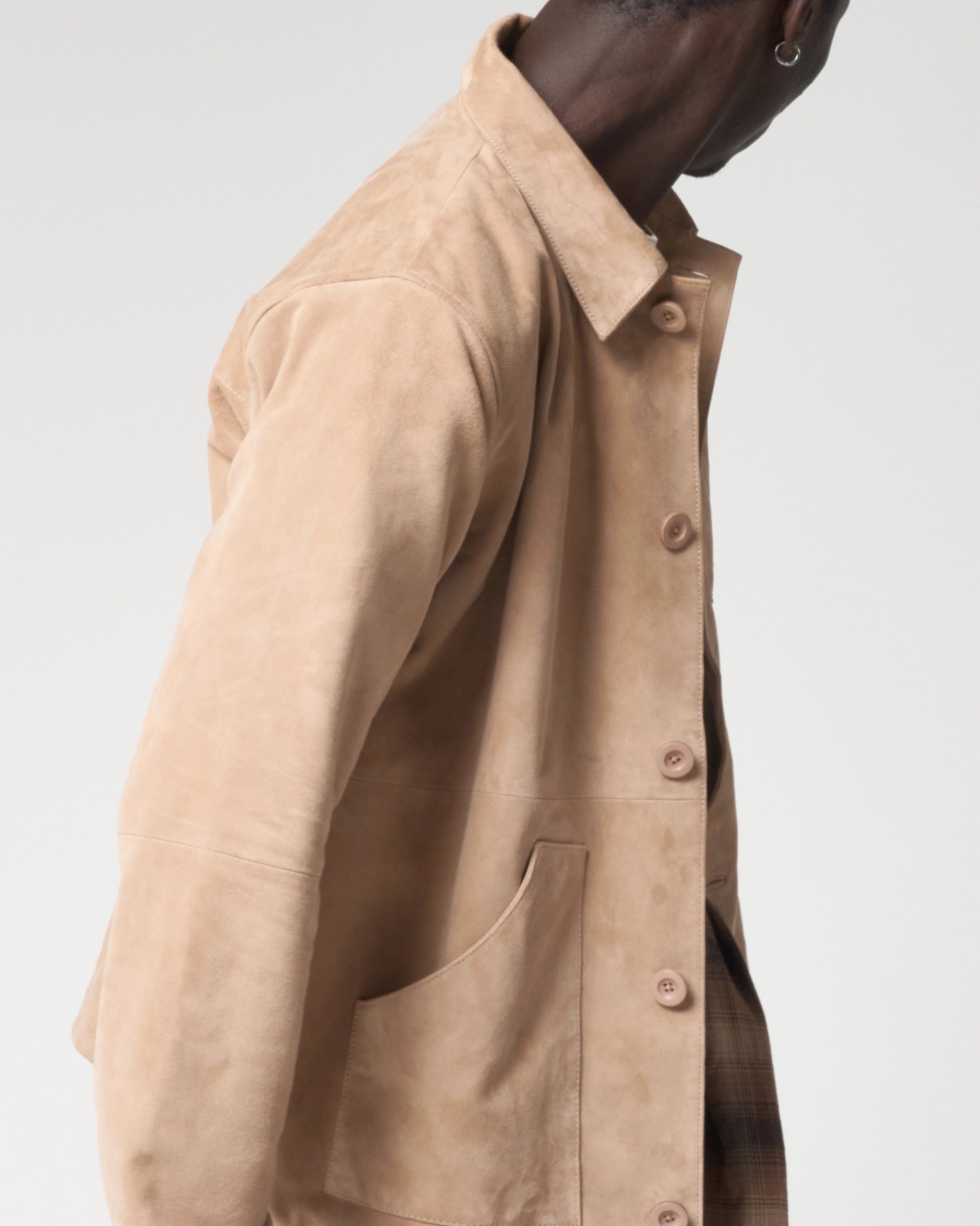 A close-up side view of a person wearing a light beige suede workwear chore jacket with a collar and large front patch pockets.
