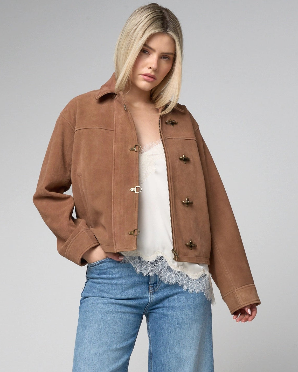 Woman wears a tan suede jacket, unzipped and open, with fireman metal clasps which line the central zipline.