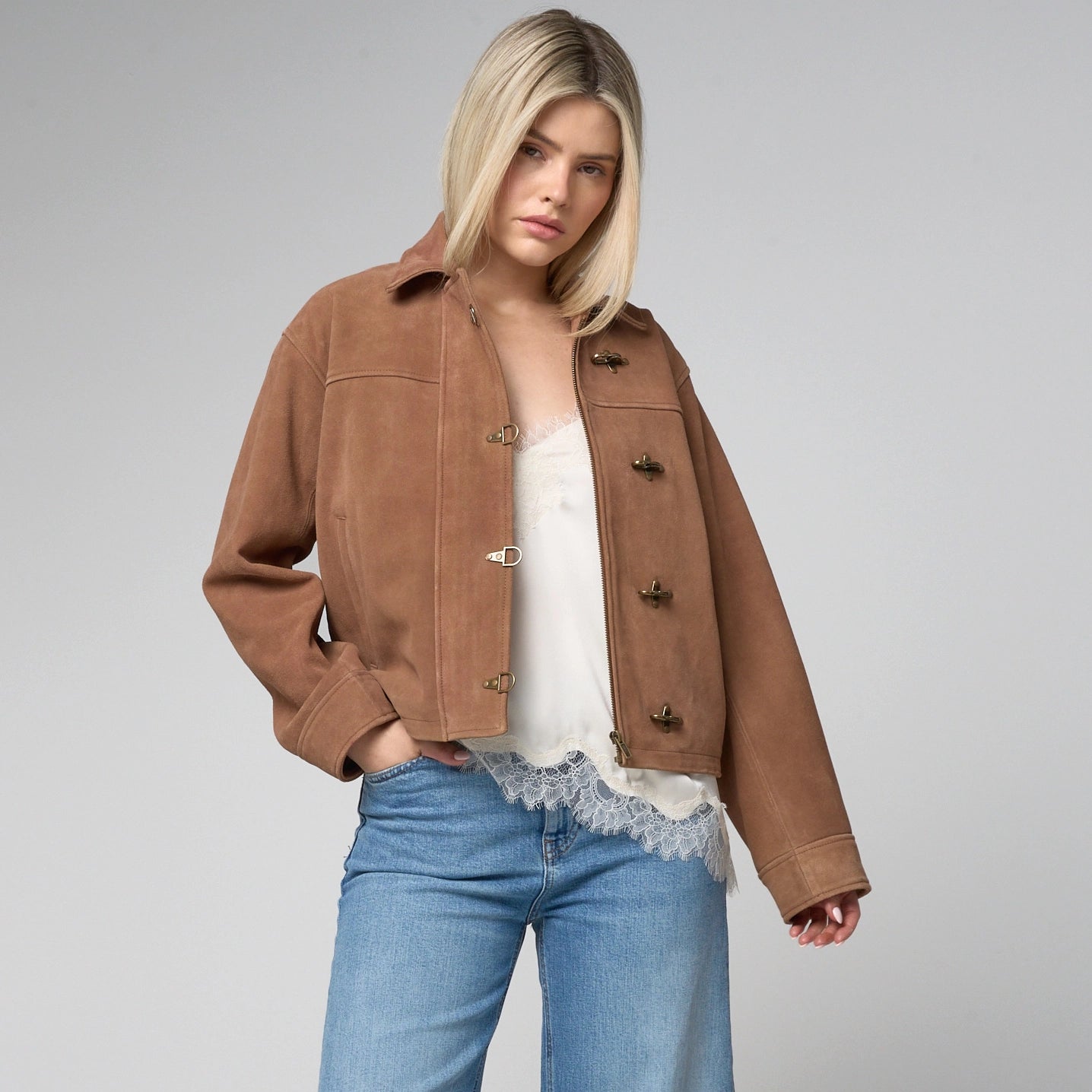 Woman wears a tan suede jacket, unzipped and open, with fireman metal clasps which line the central zipline.