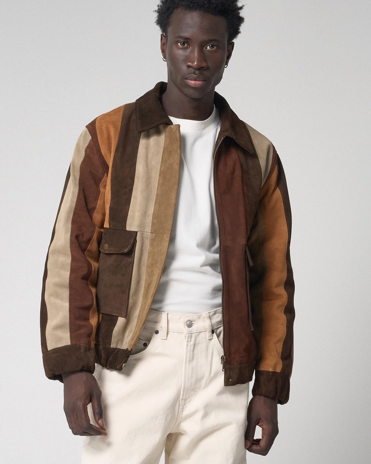 A man wearing a multi-colored patchwork jacket made of brown, tan, and beige suede panels, styled with a white t-shirt and off-white trousers against a light grey background.