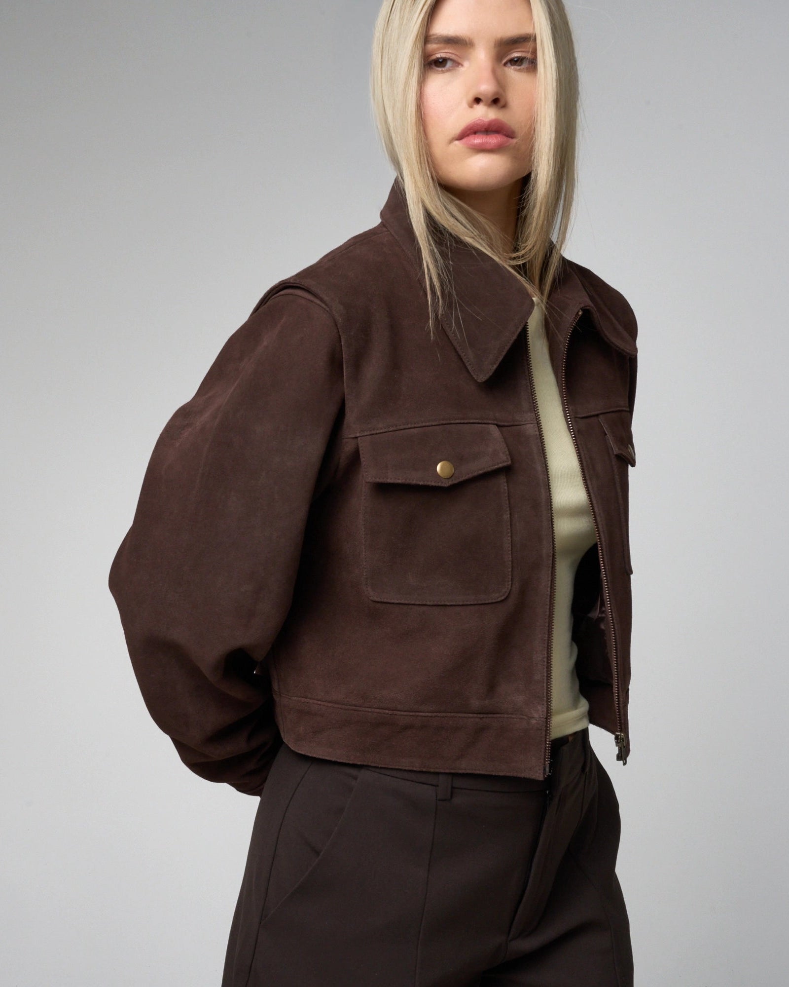 Person wearing a cropped brown suede jacket on a grey background.