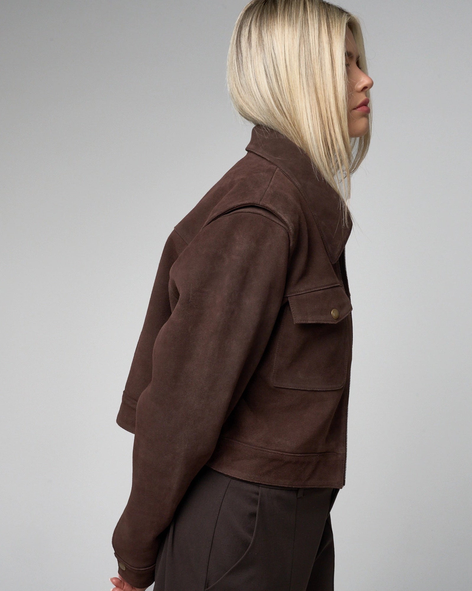 Side view of woman wearing brown suede jacket to highlight the cropped fit of the jacket which falls just above the hips when worn.