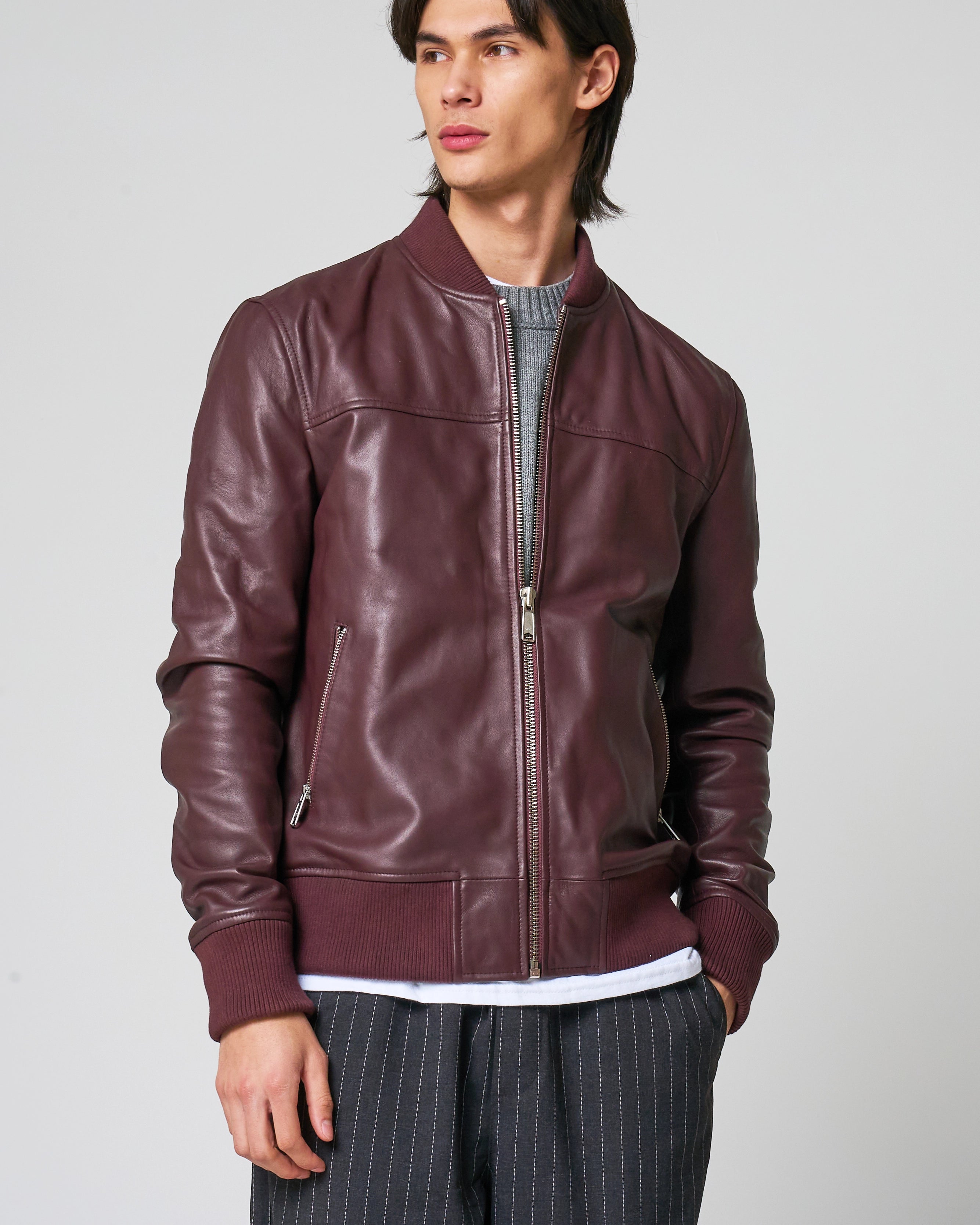 A man wearing a burgundy leather bomber jacket over a grey knit sweater and dark pinstriped trousers, photographed against a light grey studio background.