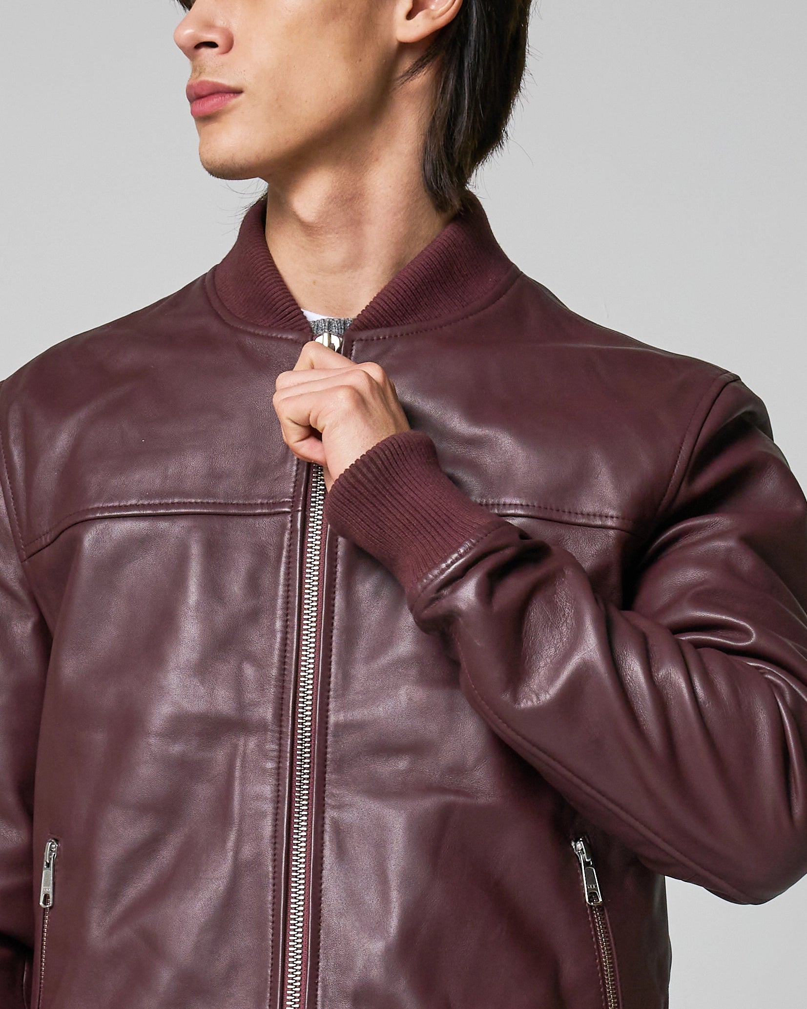 A close-up of a person wearing a burgundy leather bomber jacket, holding the zipper with one hand. The jacket has a knit rib cuff and neckline.