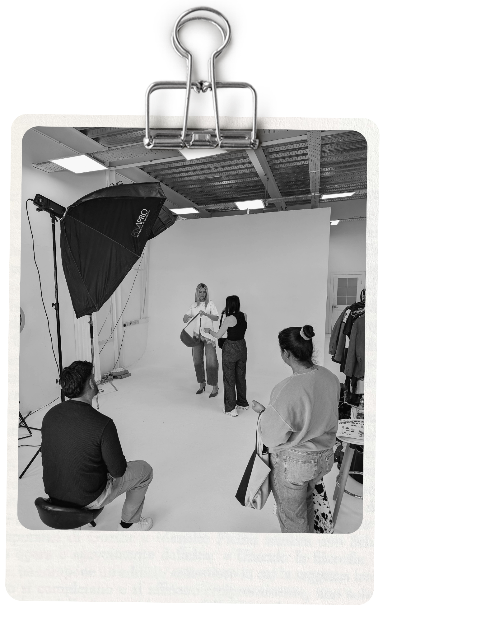 A black and white photo print clipped at the top with a metal binder clip, showing a studio fashion shoot scene with several people in a white studio space.