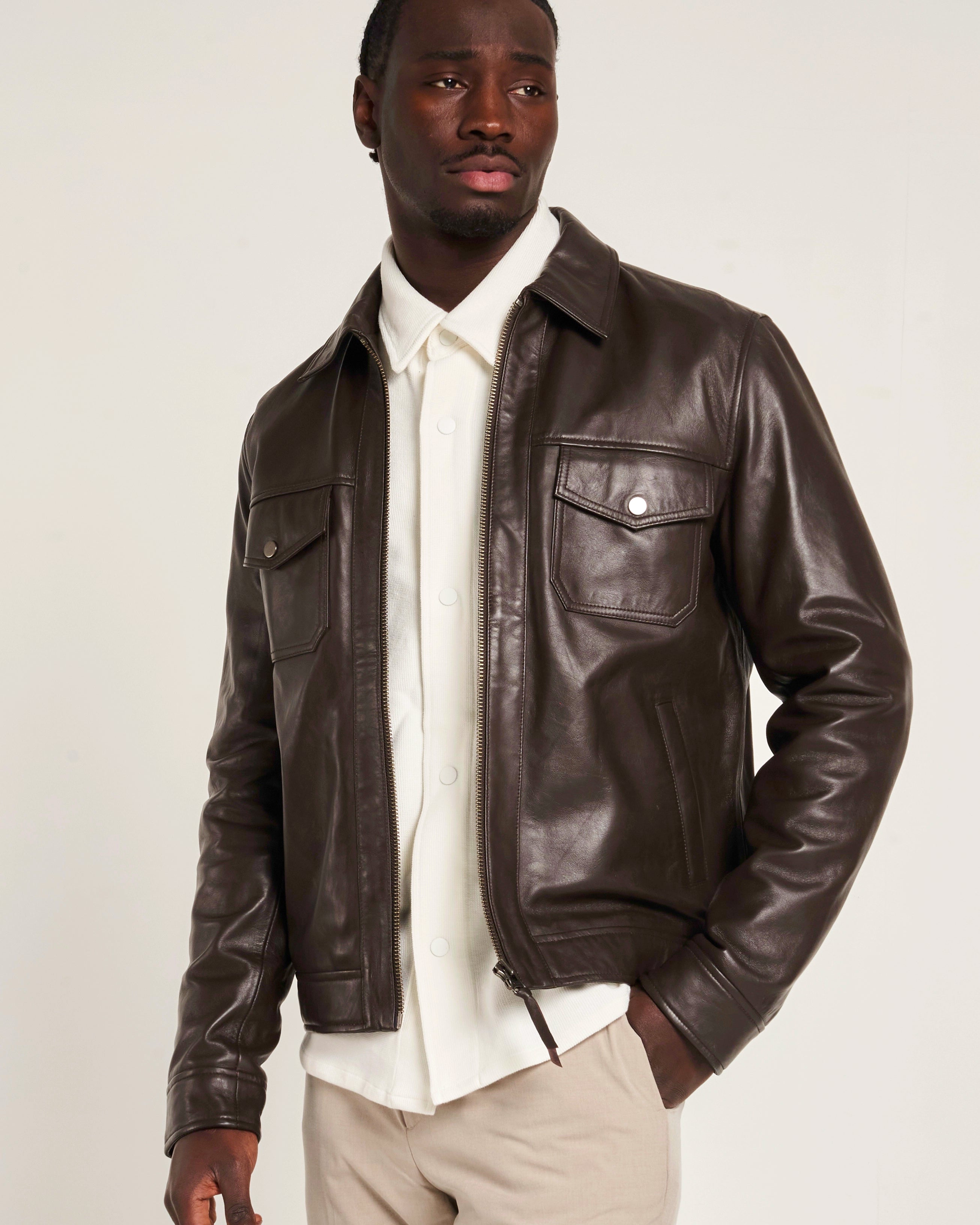 A man wearing a dark brown leather jacket over a white collared shirt and beige trousers, photographed against a plain light background. The jacket has a central zipline, lower welt pockets and upper patch pockets.