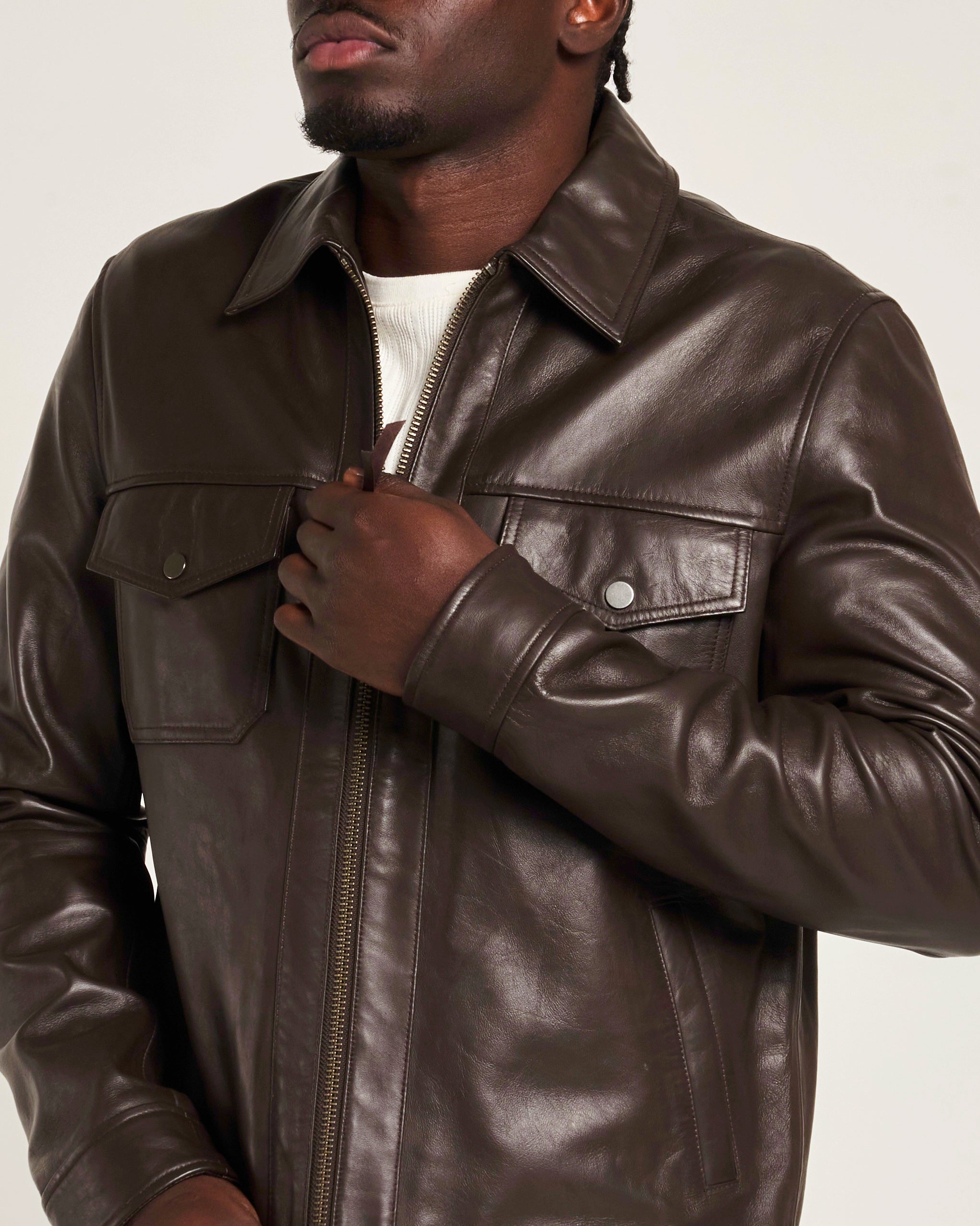 A man wearing a dark brown leather jacket with a collar, zippered front, and chest flap pockets, shown against a plain light background.