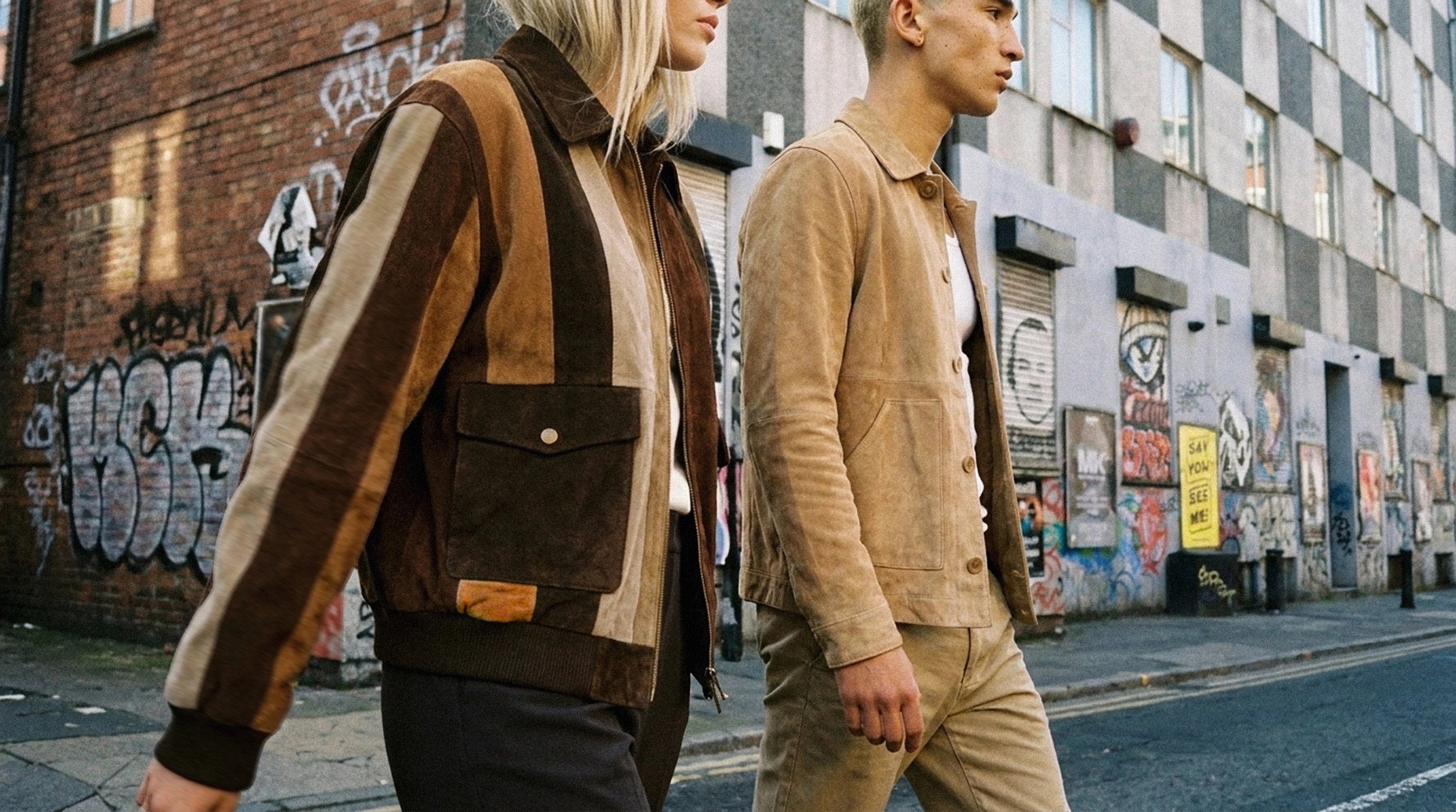 A man and a woman walking side-by-side down an urban street lined with graffiti-covered brick buildings. The woman wears a brown and tan patchwork corduroy jacket, while the man wears a tan suede jacket and beige trousers.