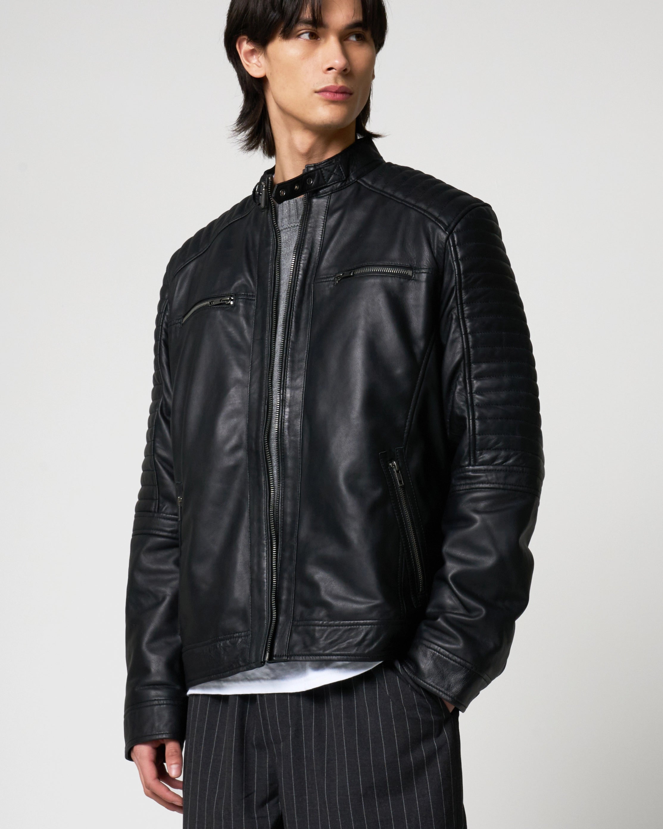 A man wearing a black leather jacket with quilted shoulder panels, zippered chest pockets, and ribbed cuffs, paired with pinstriped trousers against a light grey studio background.