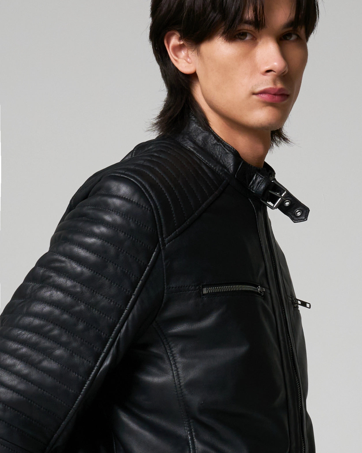 A close-up of a man wearing a black leather motorcycle jacket with quilted sleeve panels and a high collar. The collar has a buckle detail.