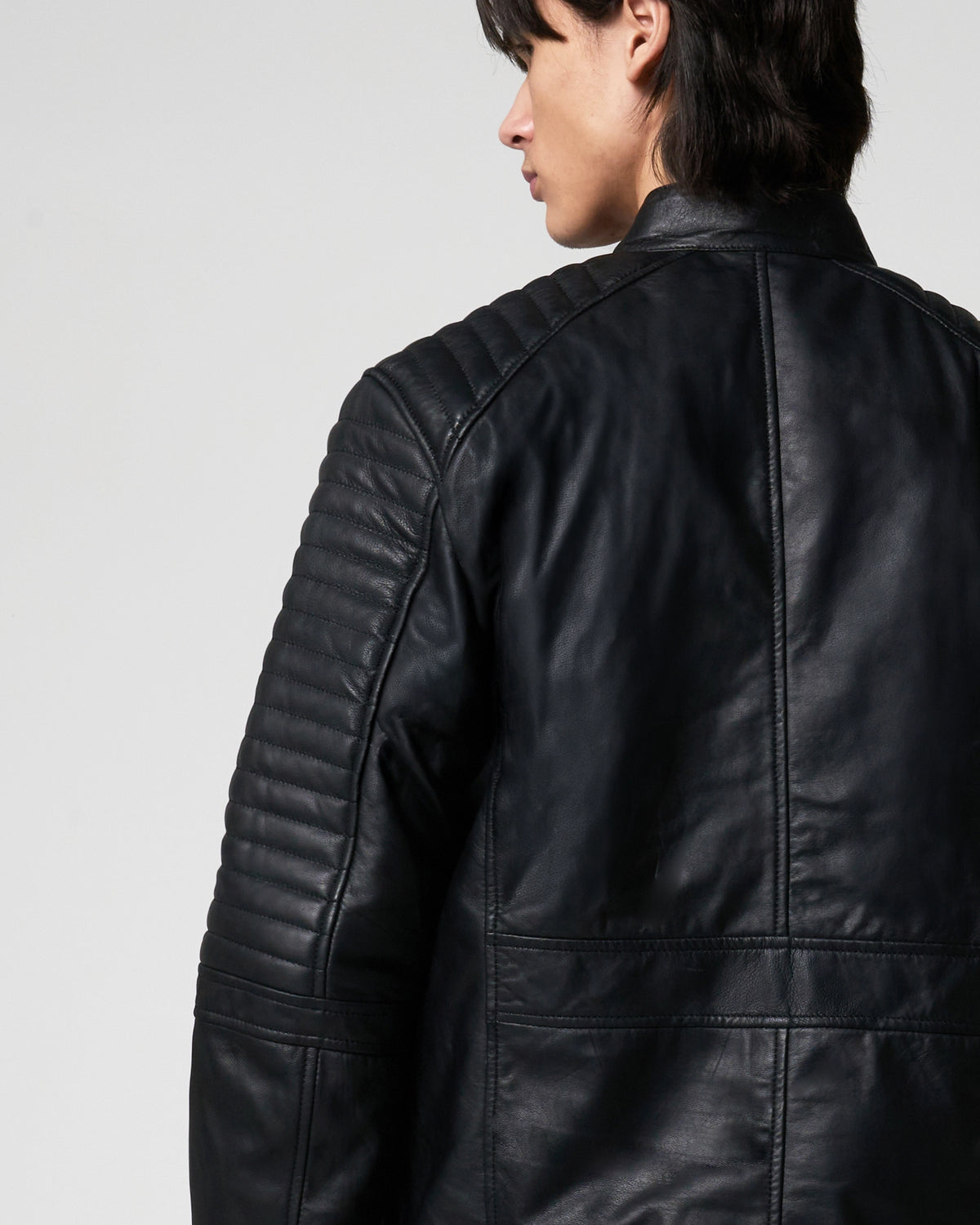 A close-up back view of a person wearing a black leather jacket with quilted shoulder panels and a high collar, photographed against a light grey studio background.