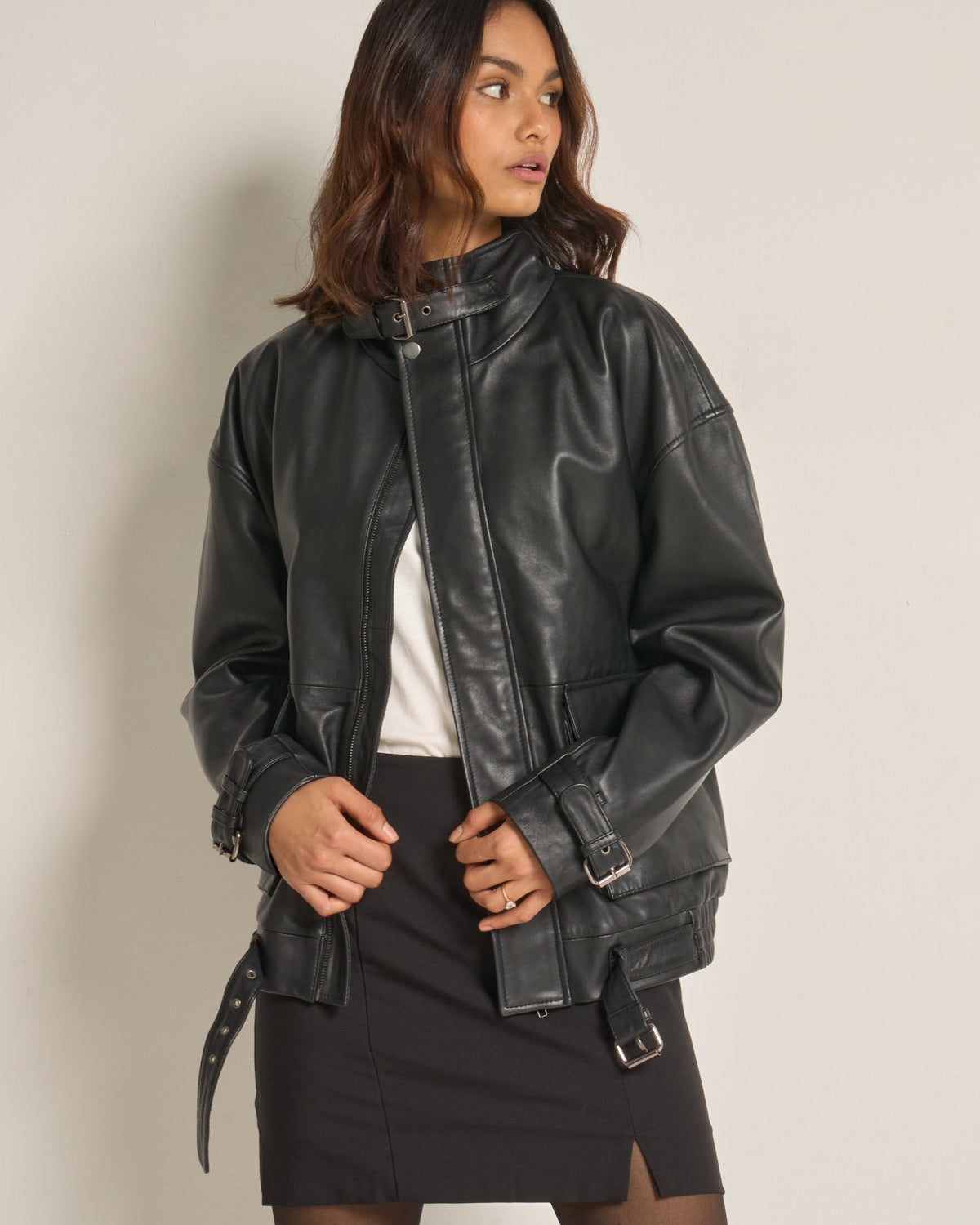 A woman wearing a black real leather moto jacket with a high collar, front zipper, and side buckle straps, styled over a white top and black skirt against a plain light grey studio background.