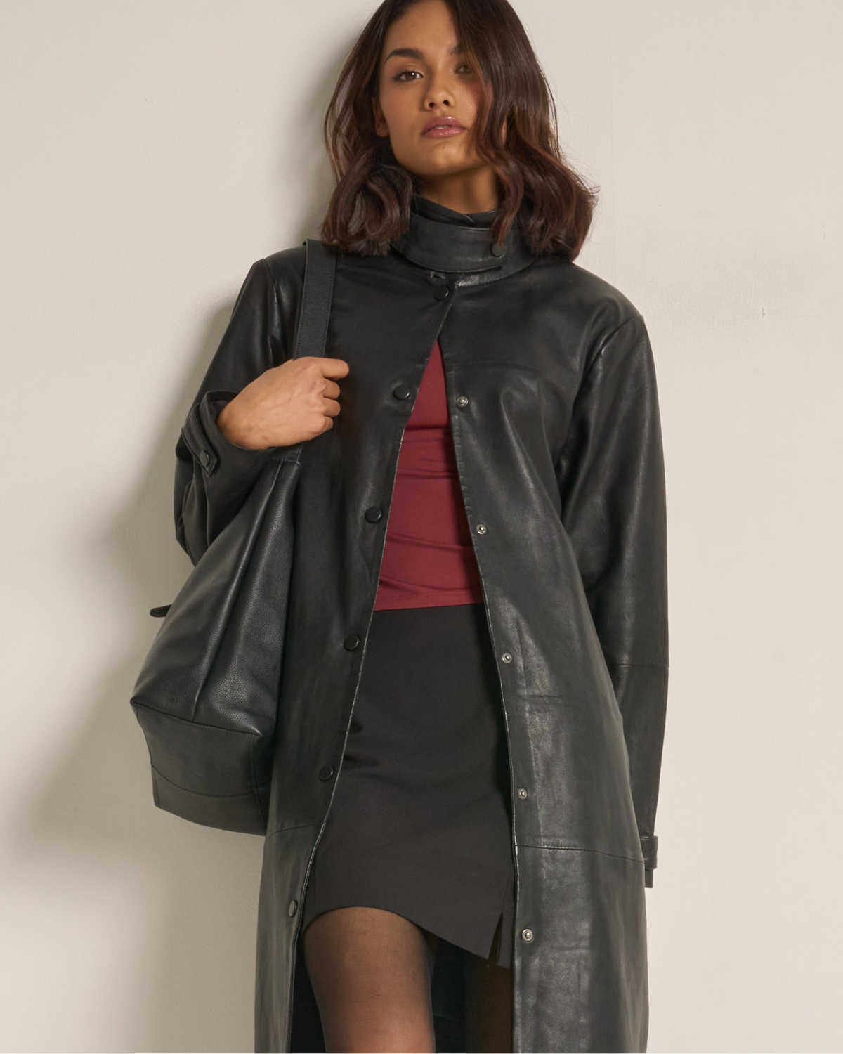 A woman wearing a long black leather coat over a dark skirt and red top, carrying a black shoulder bag against a plain off-white wall.