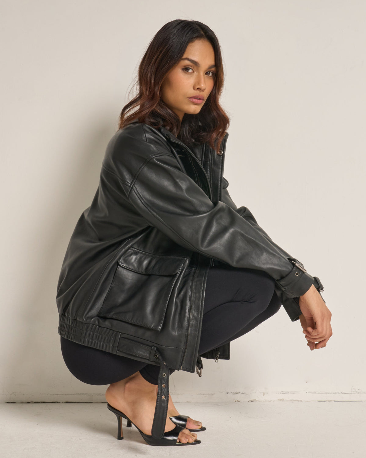A woman crouches in a studio wearing an oversized black leather jacket, black leggings, and black strappy high-heeled sandals against a plain off-white wall.