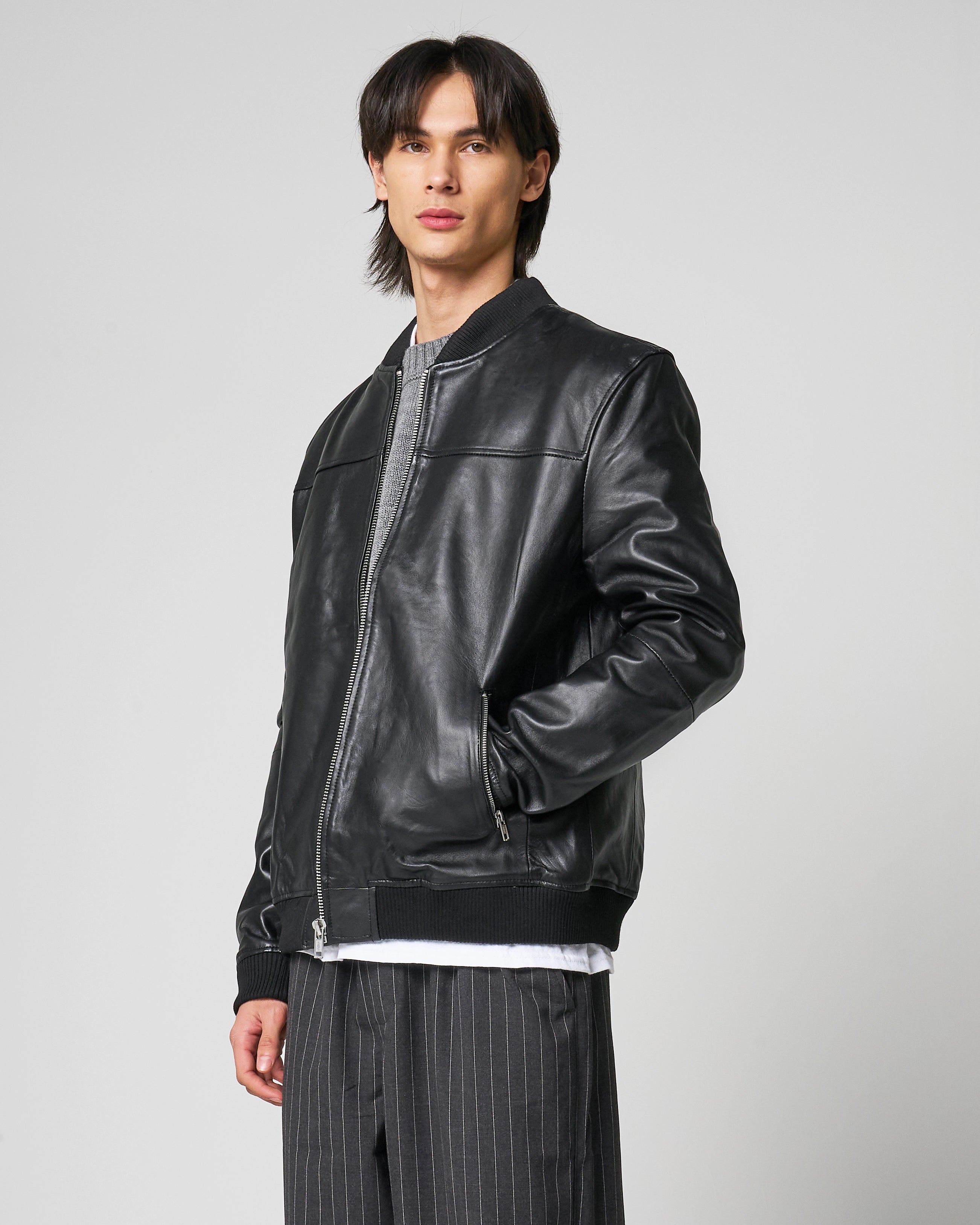 Male model wears real leather bomber jacket that is black in colour. The man puts his hand inside the lower waist pocket. Central zipline is closed half way.