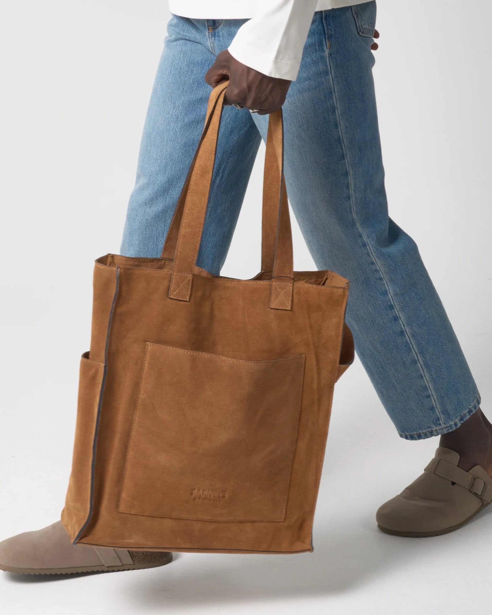 Man carries a tan suede tote back with two handles and two outer pockets.