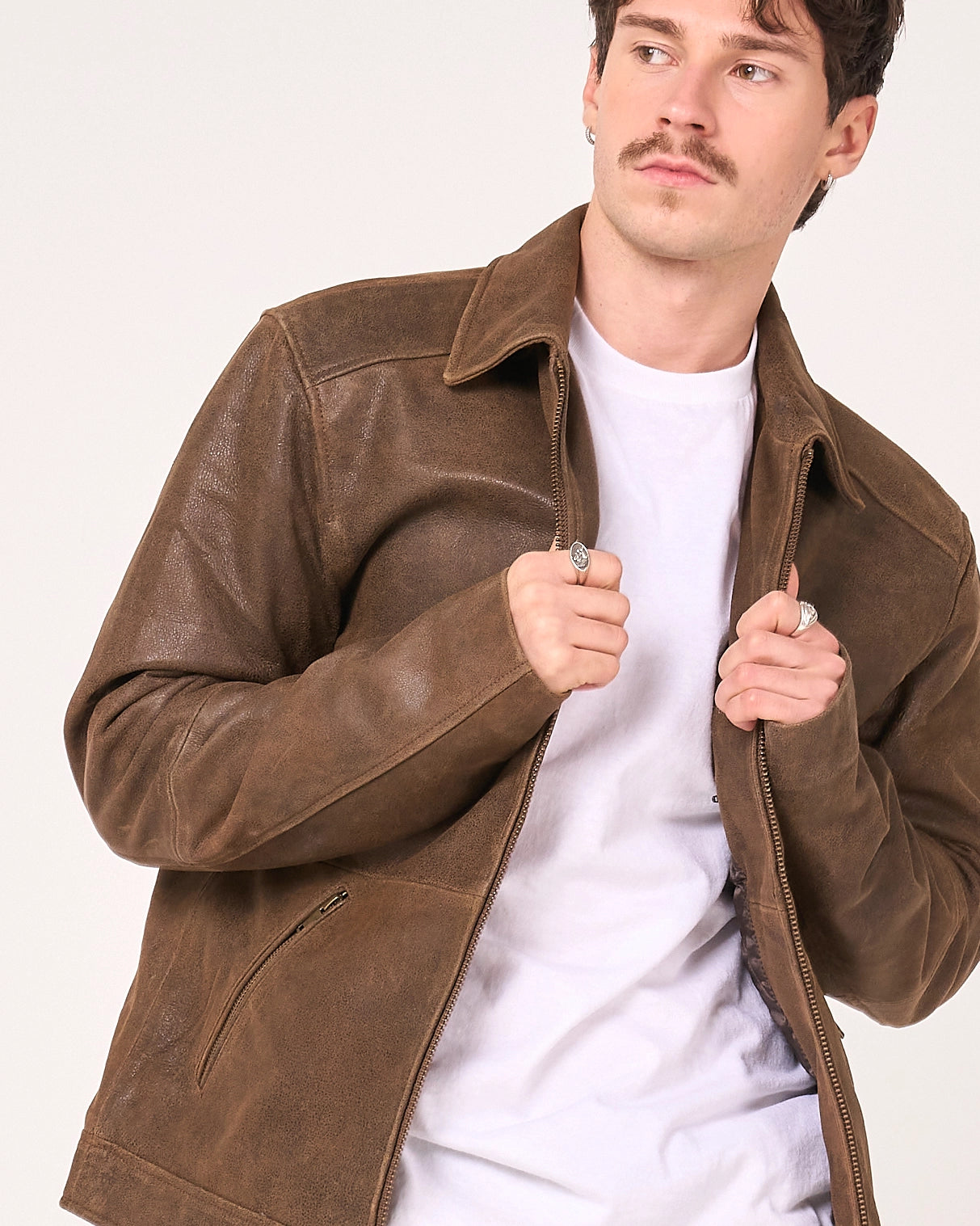 A man wearing a brown leather jacket over a white t-shirt, shown from the chest up against a plain light background.