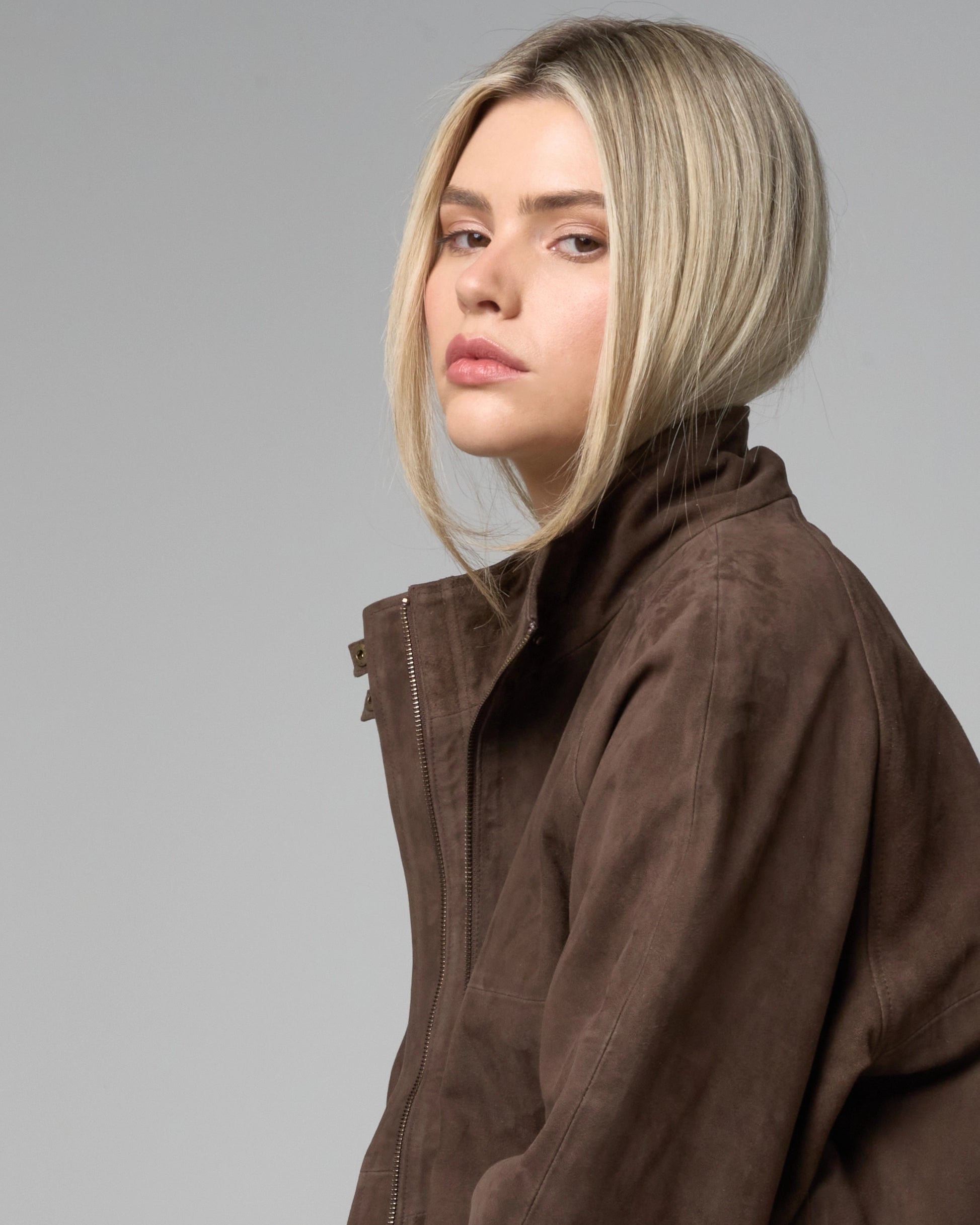 Side profile image of blond haired woman wearing a brown suede jacket with a highneck collar.