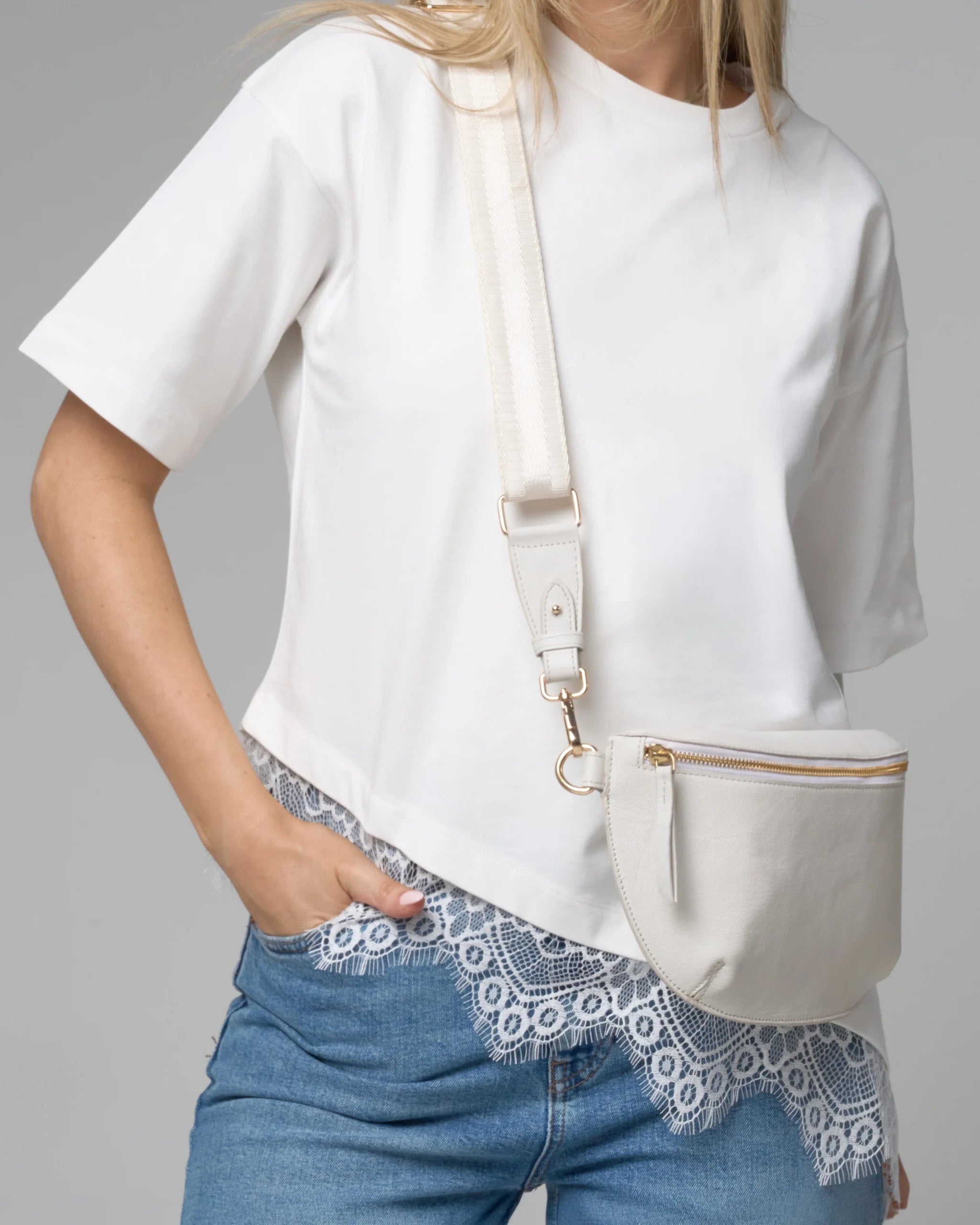 Person wearing a white t-shirt and blue jeans with a beige crossbody bag on a gray background