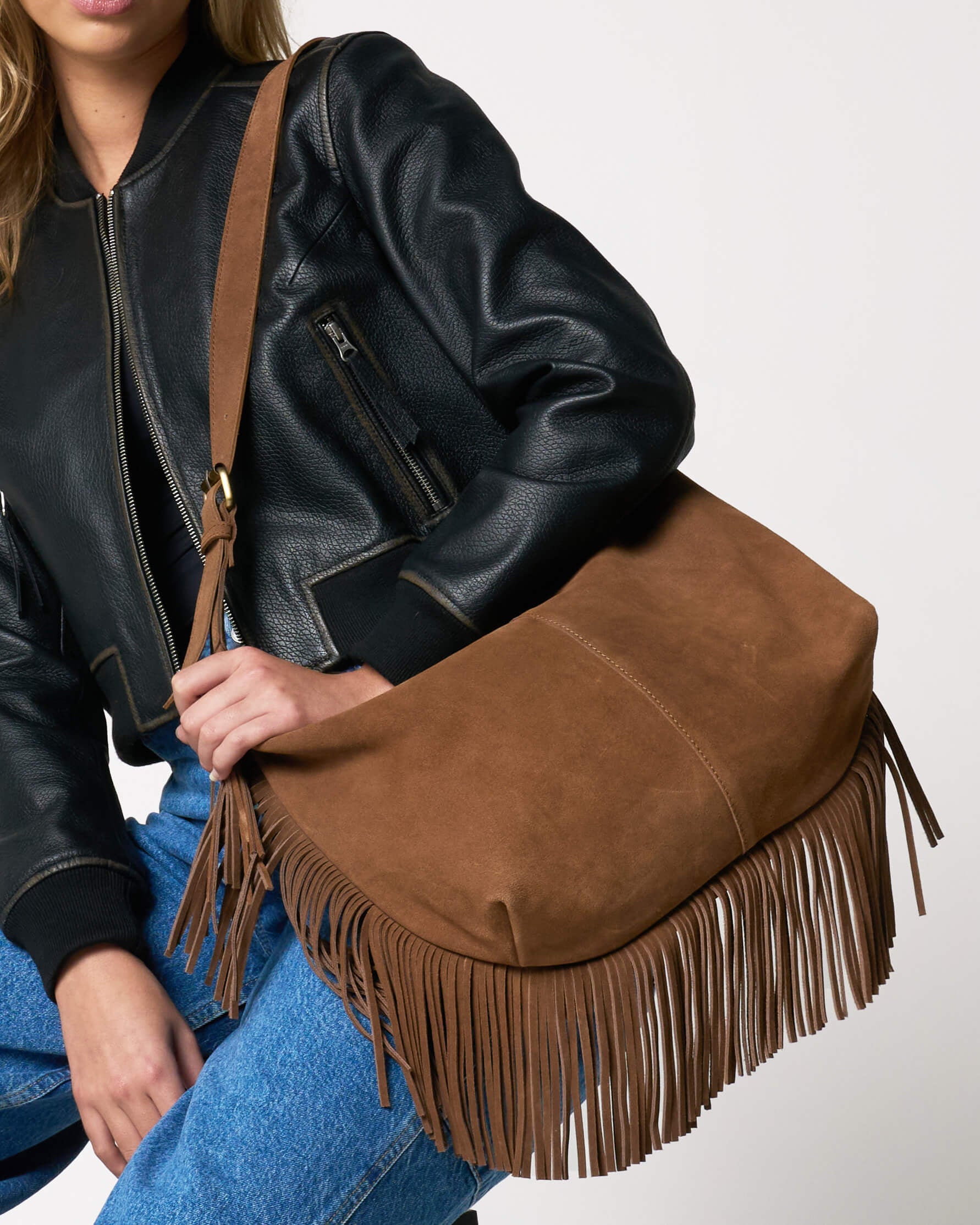 Person wearing a black leather jacket and blue jeans holding a brown suede bag with fringe detail.