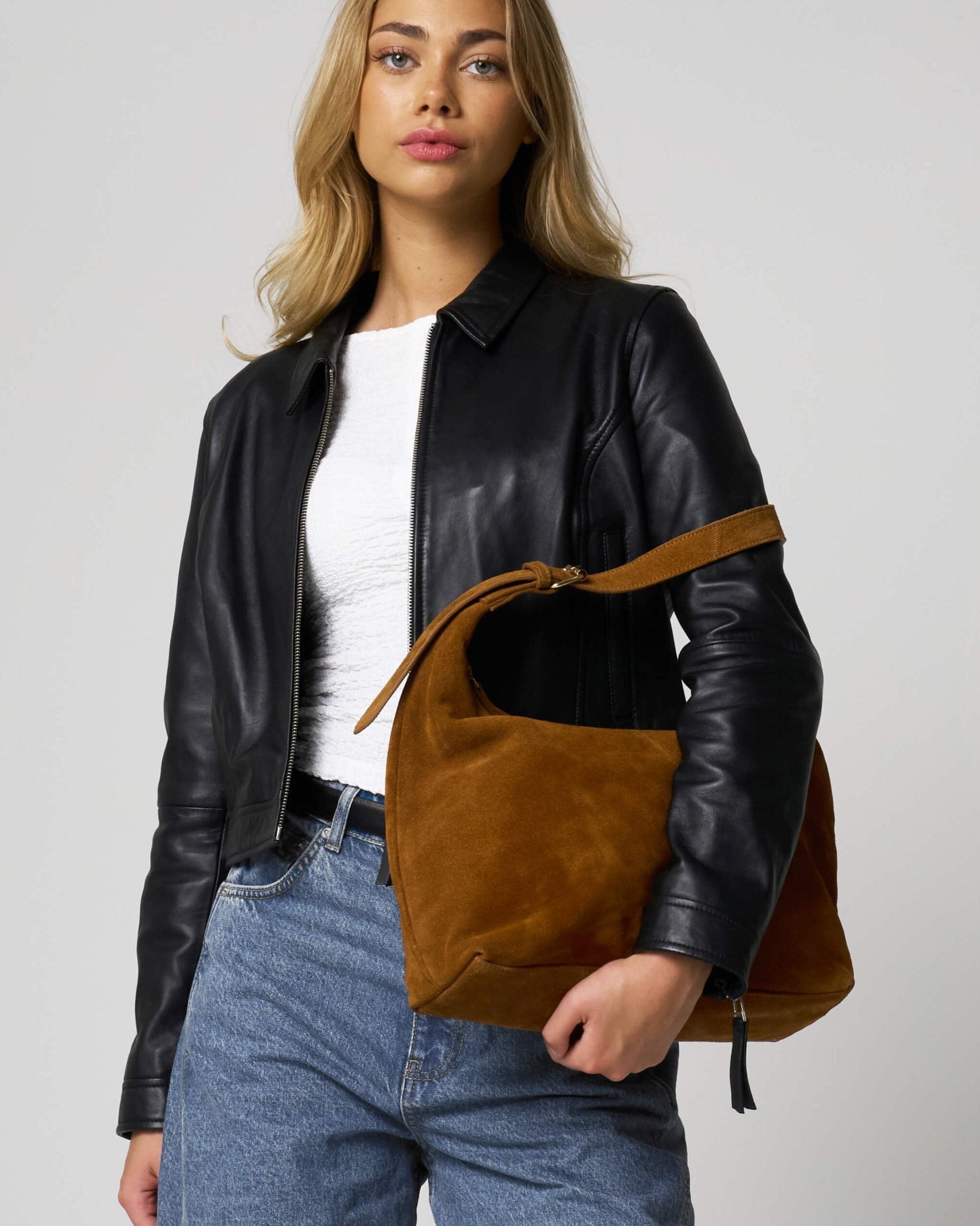 Woman wearing a black leather jacket and holding a tan suede bag against a plain background.